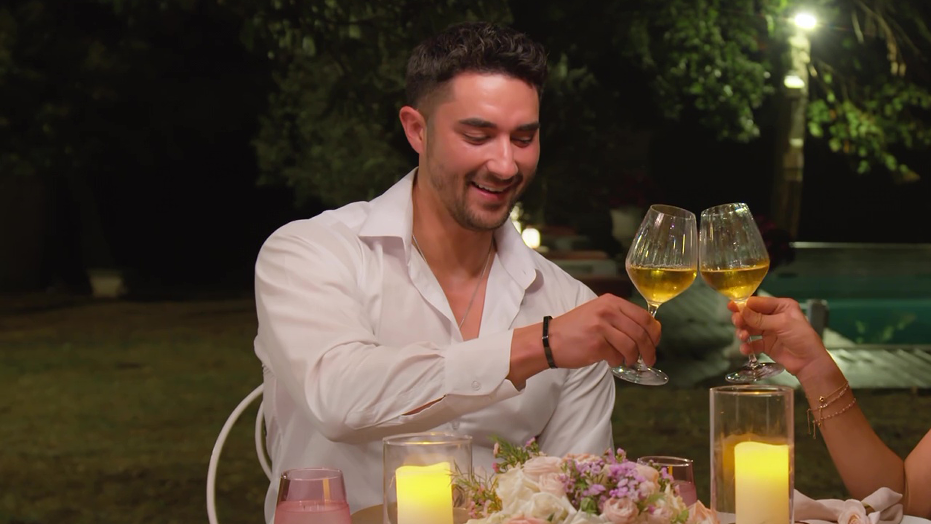 Marciano sits at a dinner table in the villa garden at night, touching his wine glass with someone else's