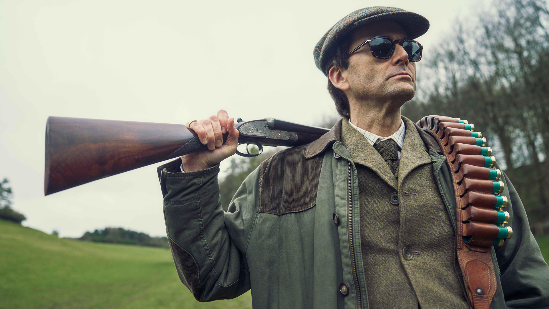 Tony Baddingham (David Tennant) com um rifle de caça pendurado no ombro esquerdo. 