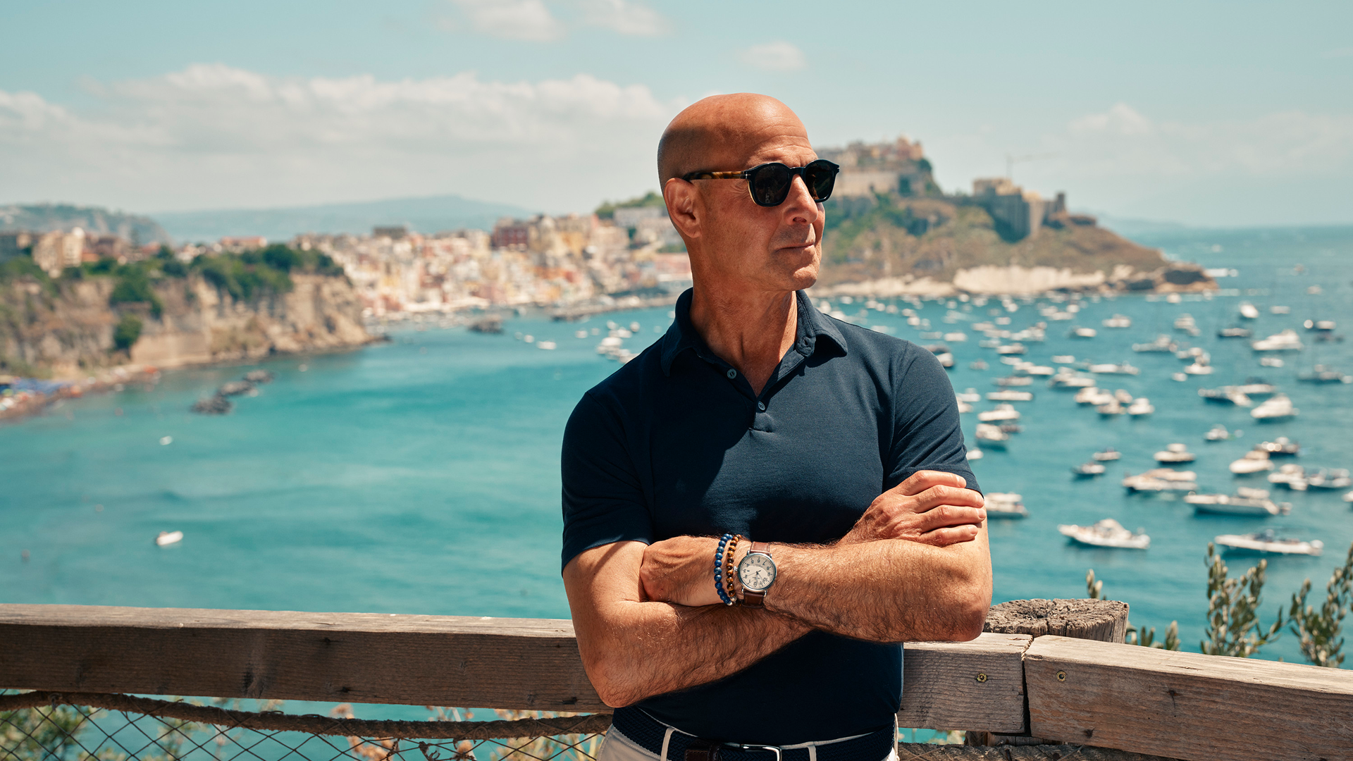 Stanley Tucci de pie frente a un paisaje costero italiano, con los brazos cruzados y anteojos de sol