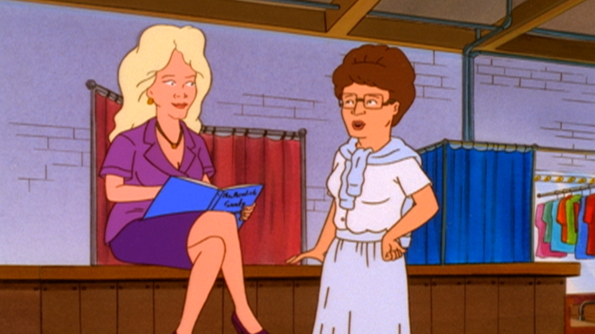 Peggy (Kathy Najimy) in a white dress talks to Nancy Gribble (Ashley Gardner) who is sat on a stage holding a clipboard.