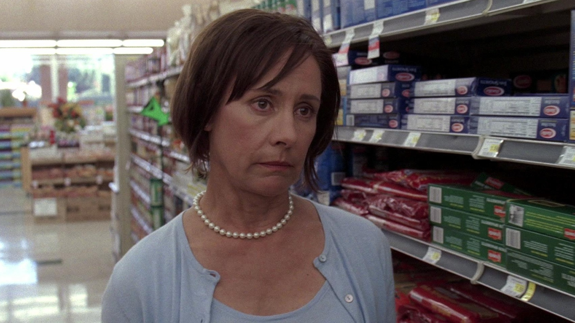 A somber Carolyn Bigsby (Laurie Metcalf) with short brown hair stands in a grocery store aisle filled with pasta boxes, wearing a pearl necklace and a light blue cardigan.