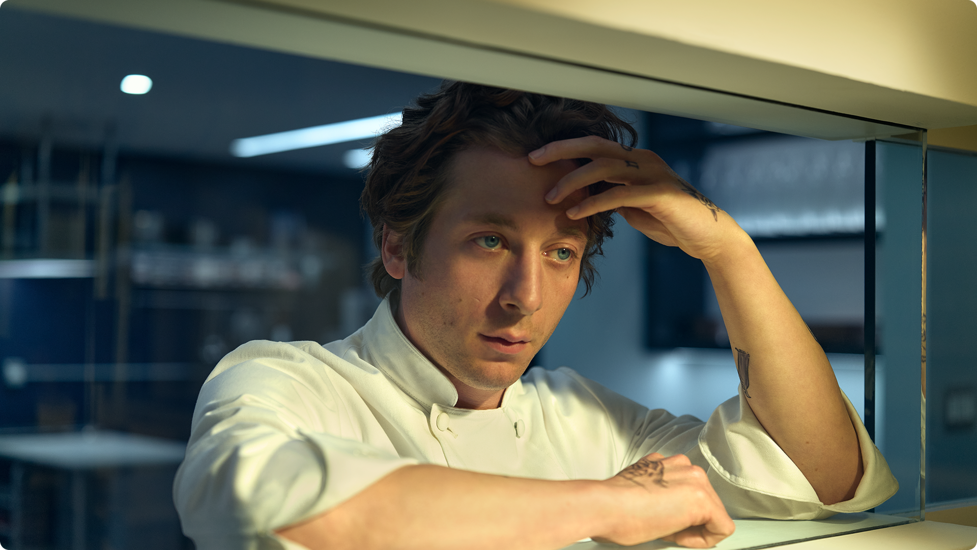A close-up shot of a pensive Carmen "Carmy" Berzatto (Jeremy Allen White) in a white chef's coat, leaning his arms against a counter in a modern restaurant kitchen while resting his forehead in his hand.