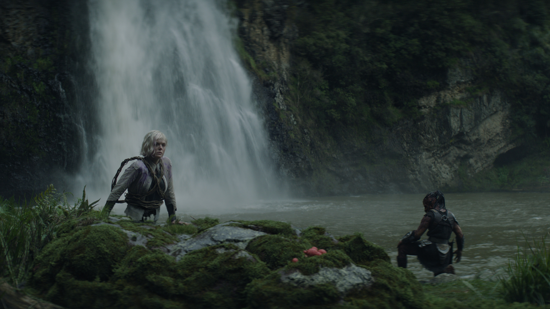 Thia (Elle Fanning) perches on rocks by a waterfall, with Dek (Dimitrius Schuster-Koloamatangi) knelt by the river nearby.