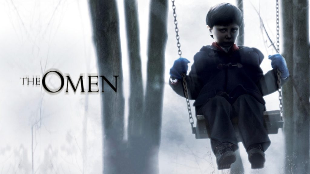 Poster for The Omen showing a pale, dark-eyed boy in a black coat sitting on a swing in a foggy forest, with the movie title displayed to the left in eerie black text.