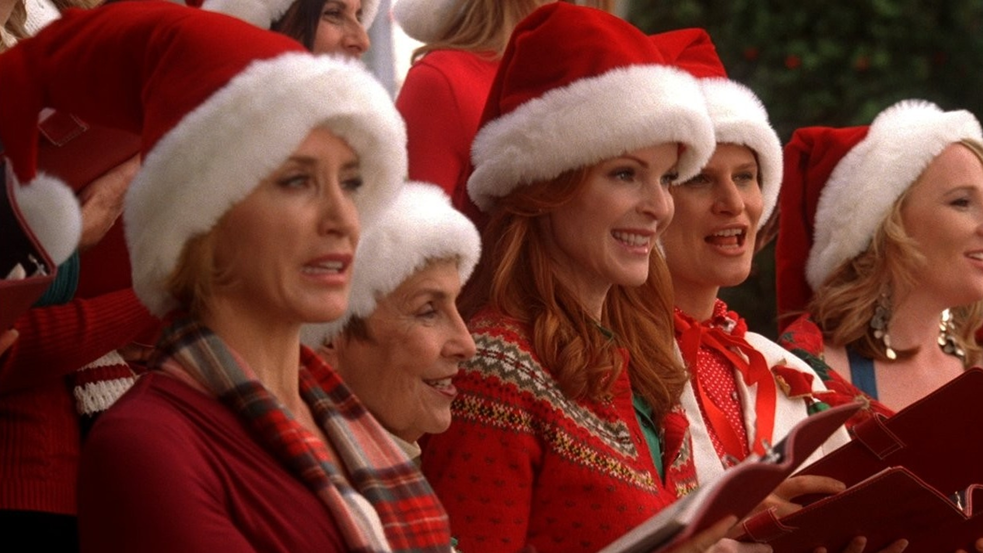 A cheerful group of Wisteria Lane residents, including Lynette Scavo (Felicity Huffman) and Bree Van de Kamp (Marcia Cross), sing holiday music while dressed in festive red sweaters and plush Santa hats.