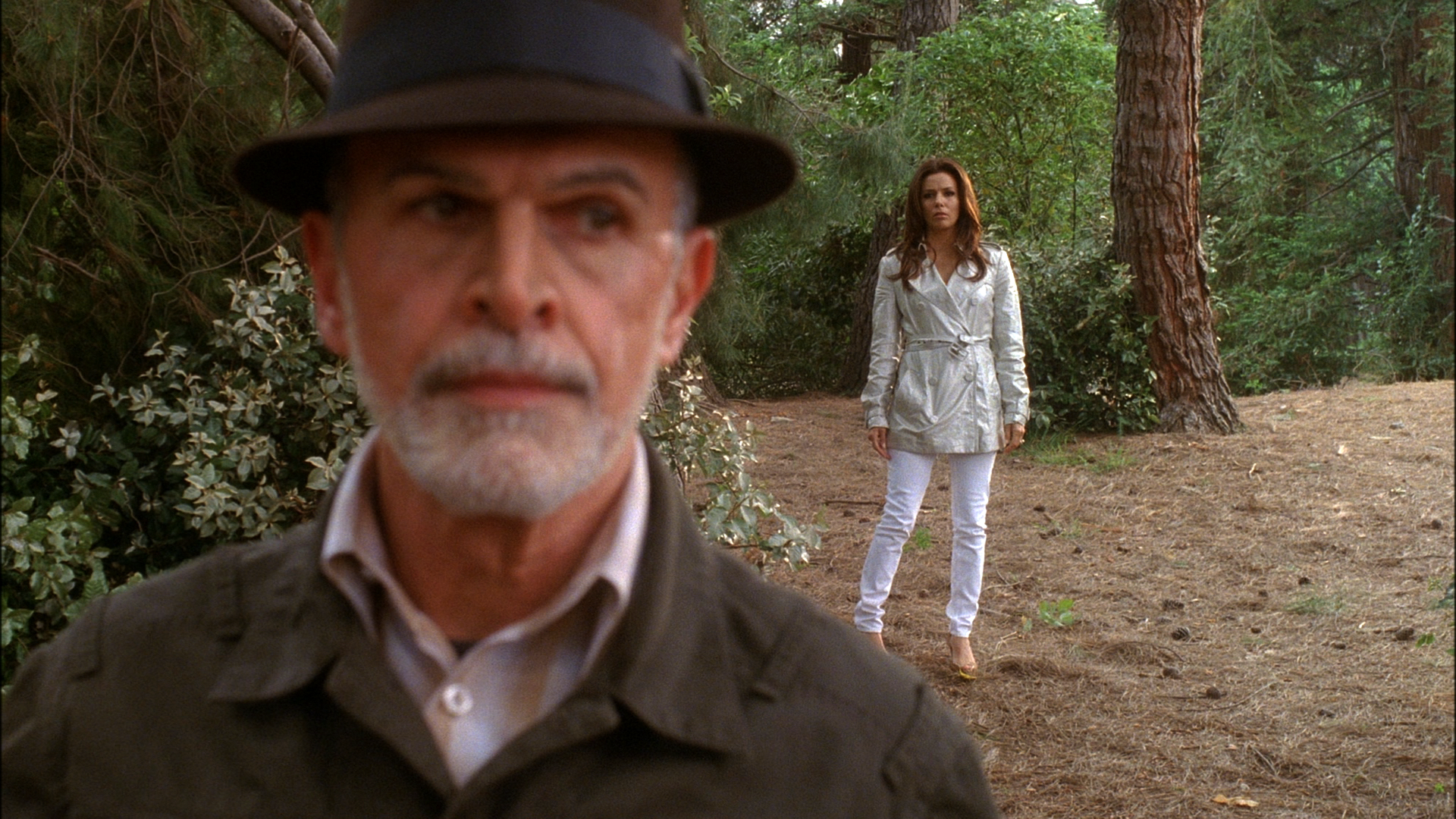 A blurred Alejandro Perez (Tony Plana) wearing a dark fedora dominates the foreground, while a tense Gabrielle Solis (Eva Longoria) stands watching him from a distance in a pine forest.