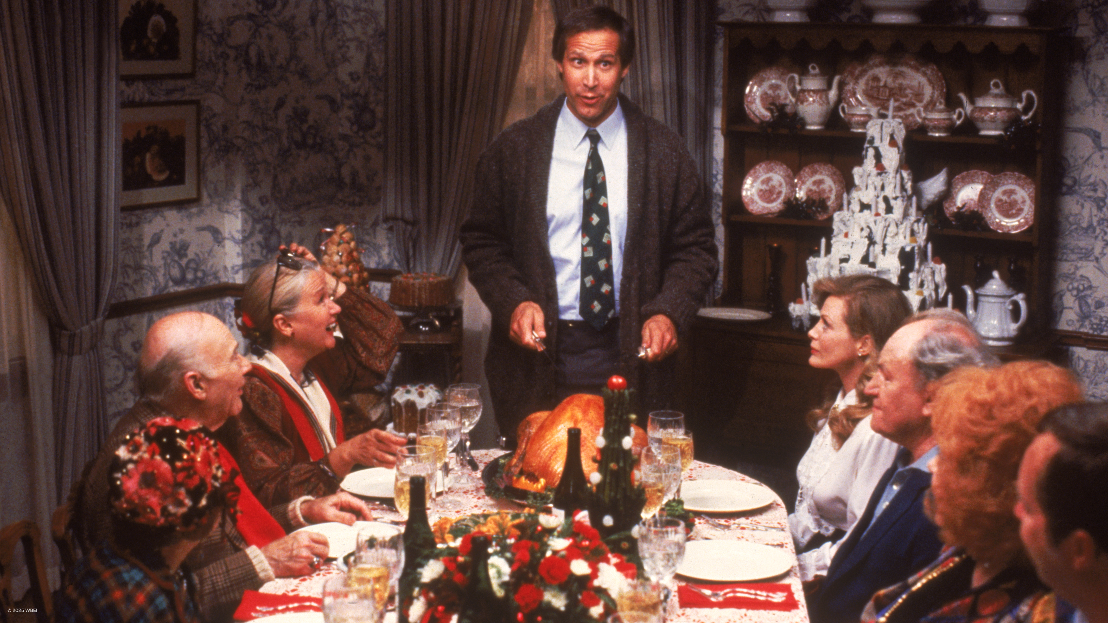 Clark Griswold (Chevy Chase) stands at the head of a dining table decorated for Christmas, preparing to carve a turkey.
