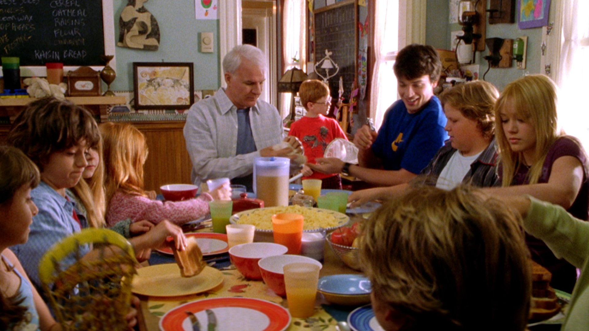 A hectic family breakfast scene from Cheaper by the Dozen shows Tom Baker (Steve Martin) sitting at a crowded dining table full of food and colorful cups, surrounded by several of his twelve children including Charlie (Tom Welling) and Lorraine (Hilary Duff).
