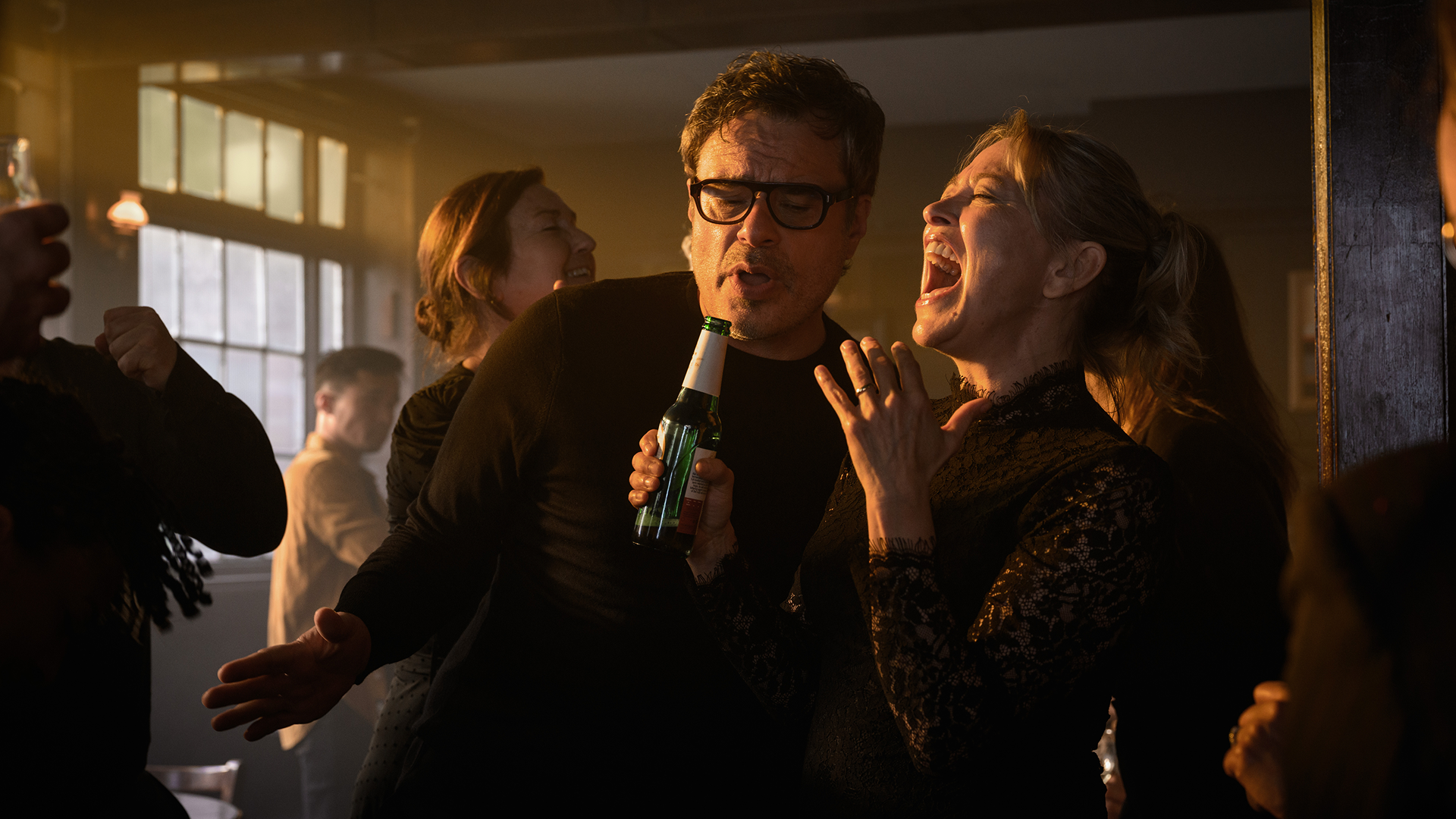 Steve (Jemaine Clement) and Alice (Nicola Walker) sing passionately in a crowded, dimly lit pub while Steve holds a green beer bottle like a microphone in a scene from Alice and Steve.