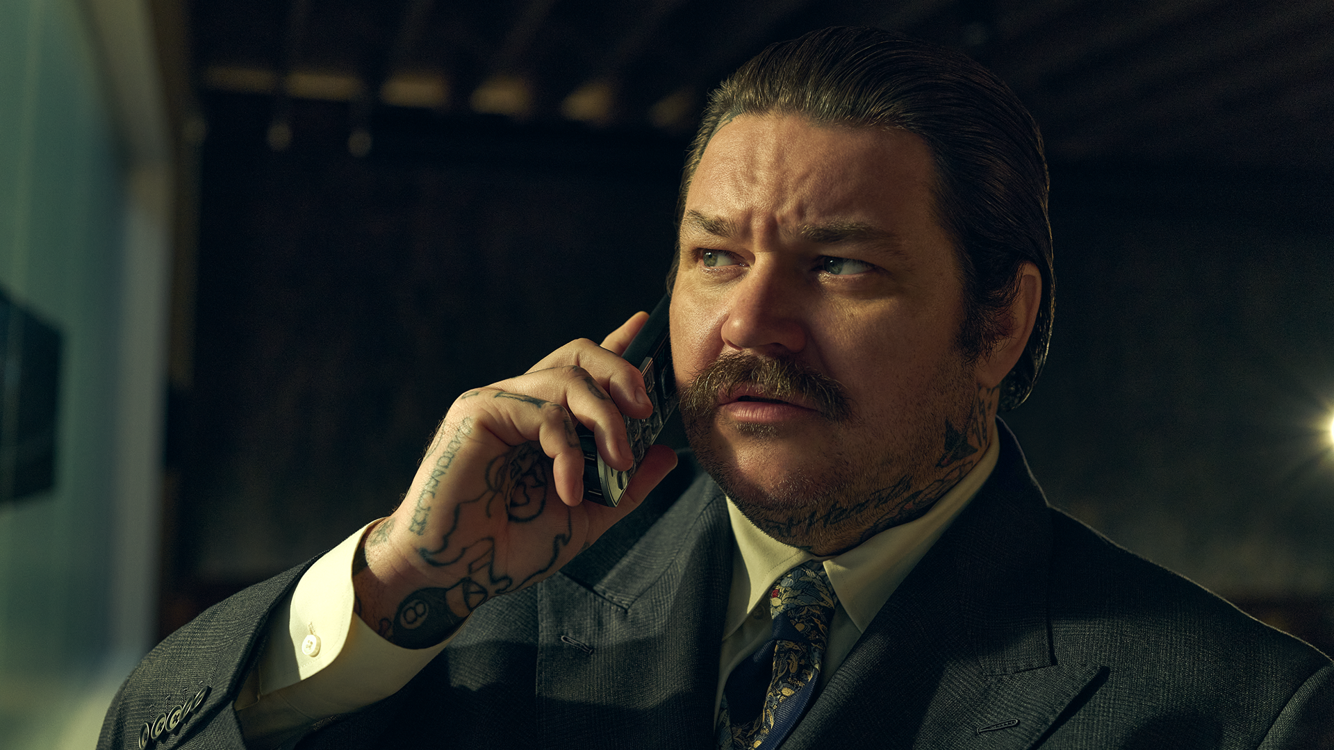 A close-up shot of Neil Fak (Matty Matheson) with slicked-back hair and a mustache, wearing a patterned tie and a charcoal gray suit jacket while holding a mobile phone to his ear with his heavily tattooed right hand.