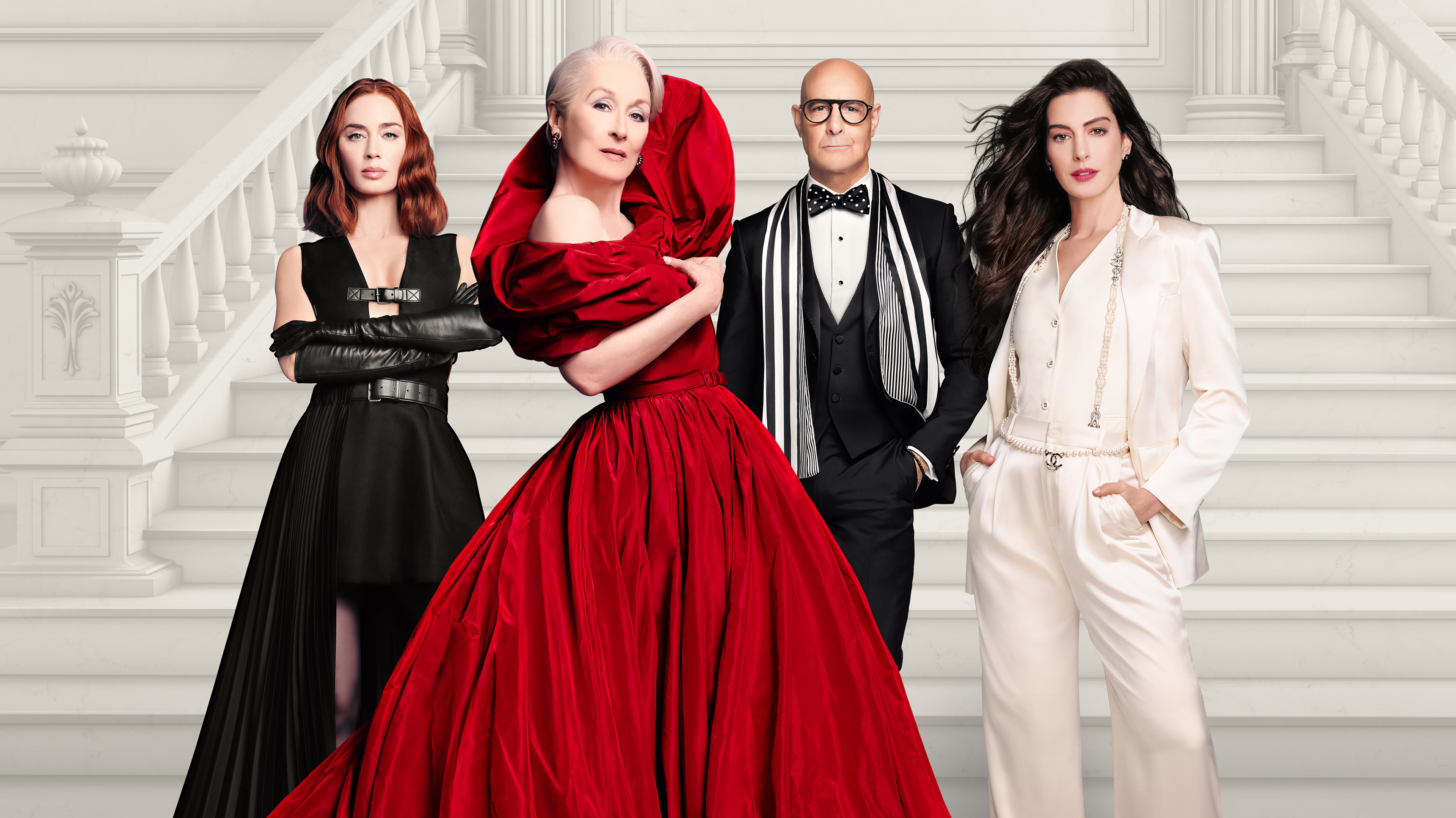 Four actors from the movie The Devil Wears Prada—Emily Blunt, Meryl Streep, Stanley Tucci, and Anne Hathaway—pose together in high-fashion black, red, and white outfits on a grand marble staircase.