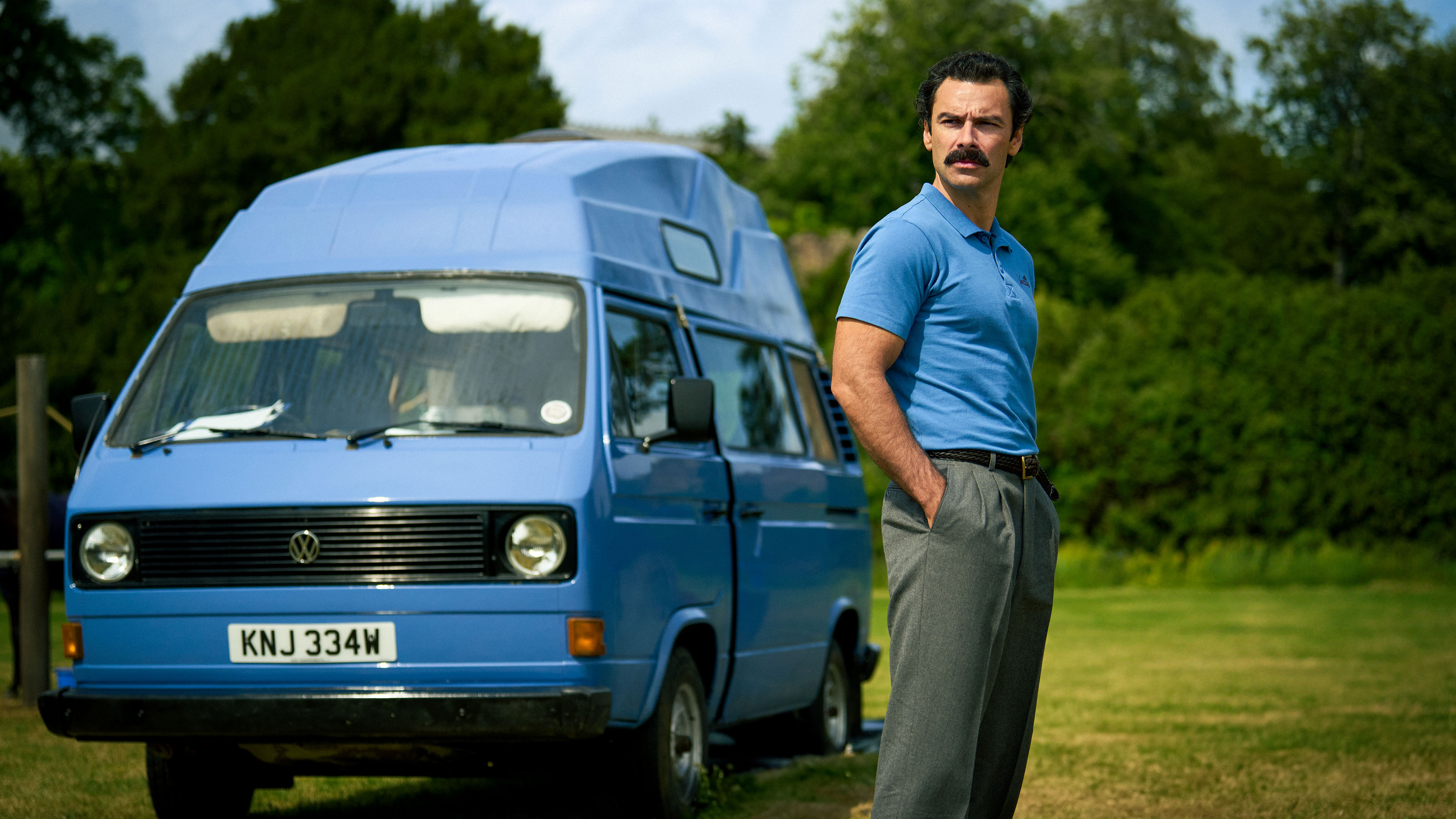 Declan O’Hara (Aidan Turner) stands beside a blue Volkswagen camper van outdoors in Rivals season 2.