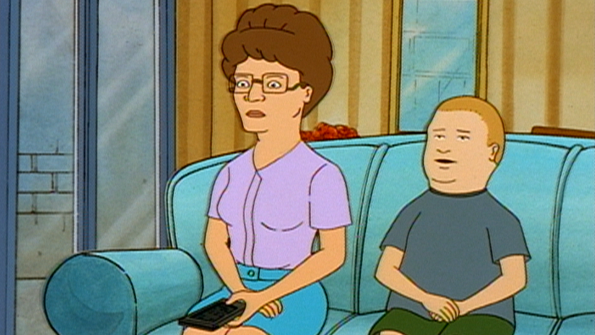 Peggy (Kathy Najimy) sits on the sofa holding a remote beside Bobby (Pamela Adlon).