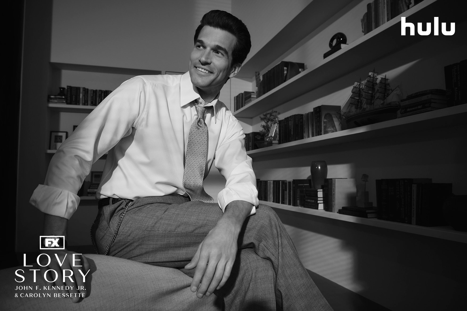 A smiling John F. Kennedy Jr. (Paul Kelly) sitting in an office in a black and white set photo from FX's Love Story.