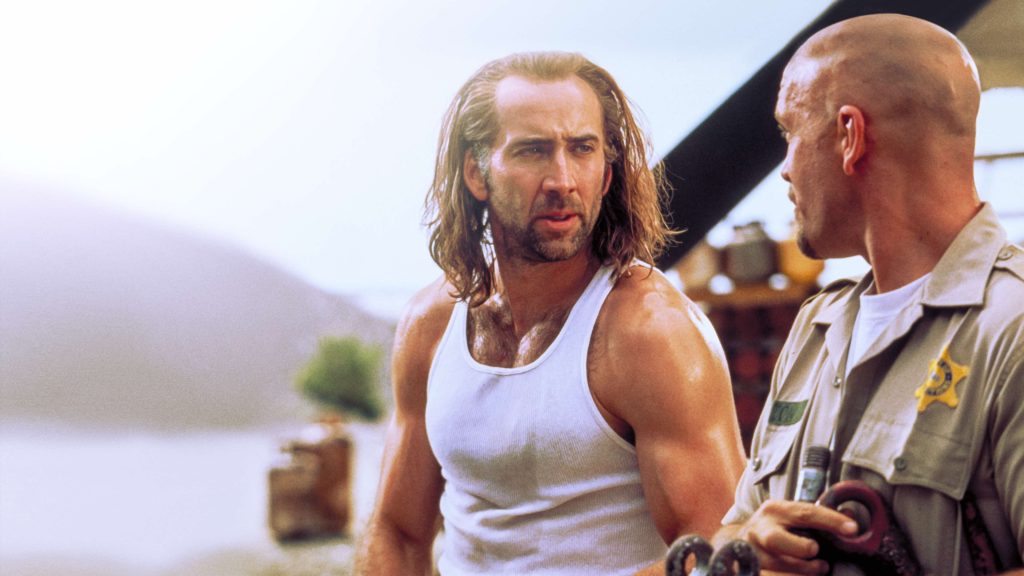 Nicolas Cage as Cameron Poe in Con Air, wearing a white tank top and long hair while talking to a guard.
