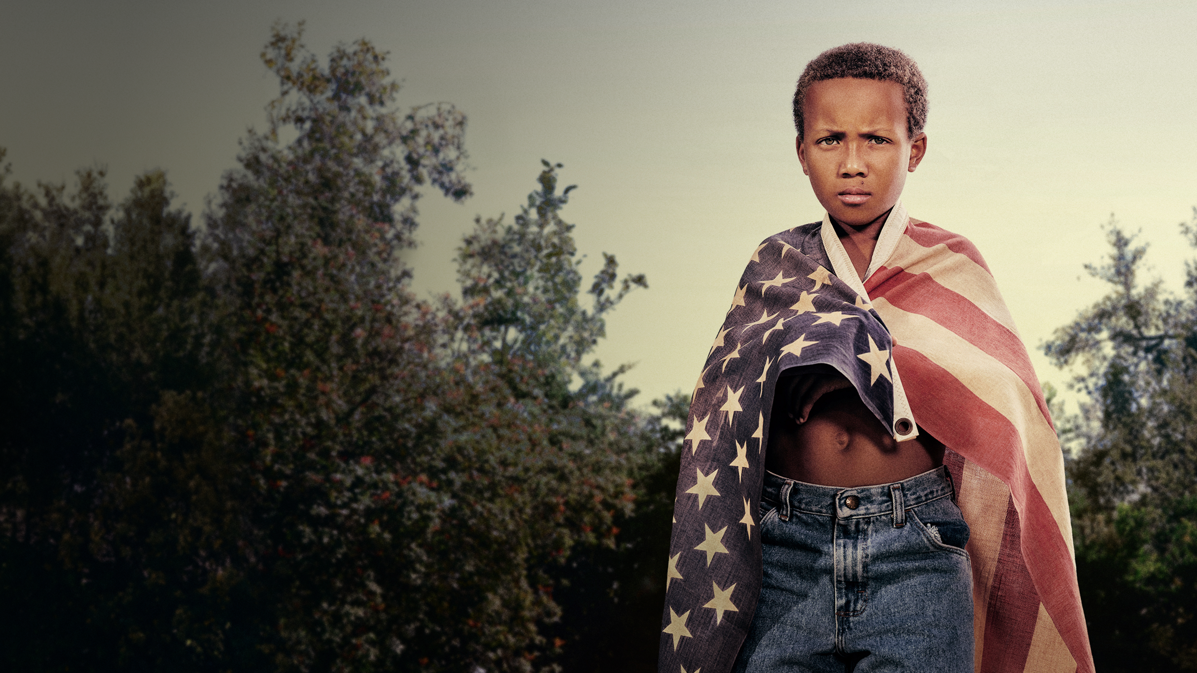 A man with locs holding a child in the air, an American flag in the background