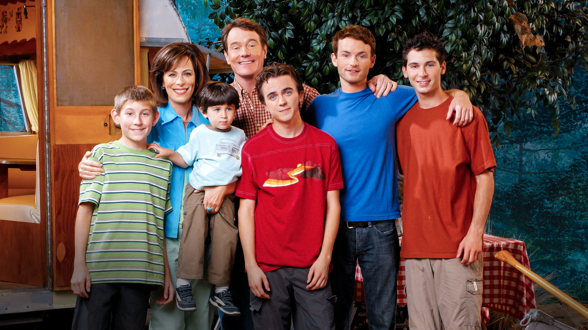 A cast promotional photo for the sitcom Malcolm in the Middle shows Hal (Bryan Cranston) and Lois (Jane Kaczmarek) standing with their five sons, Dewey (Erik Per Sullivan), Jamie (James and Lukas Rodriguez), Malcolm (Frankie Muniz), Francis (Christopher Masterson), and Reese (Justin Berfield), smiling together in front of an outdoor camper trailer and picnic setup.