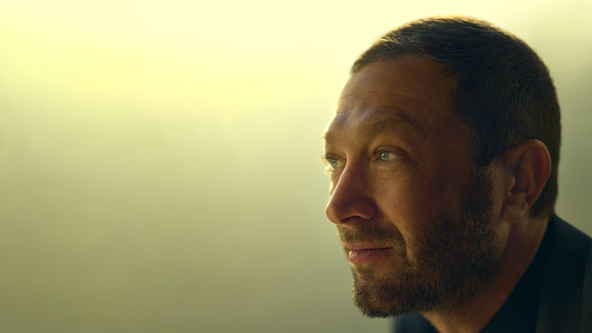 A profile close-up shot of a thoughtful Richard "Richie" Jerimovich (Ebon Moss-Bachrach) looking off into the distance against a soft, bright yellow background.