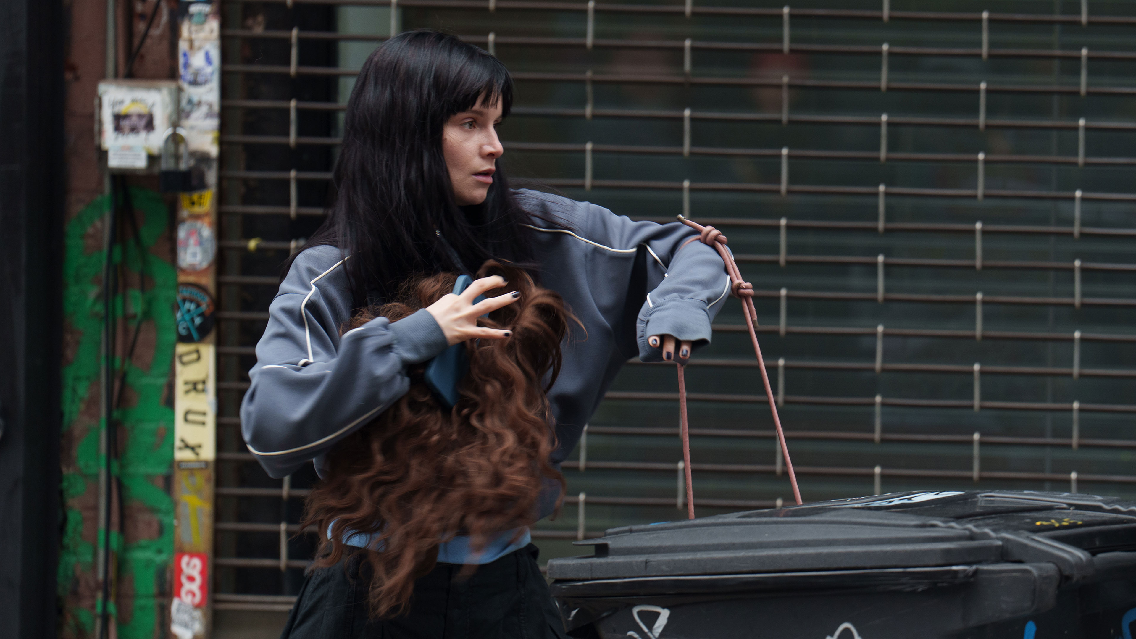Catherine holds a long wig beside rubbish bins in a city street.