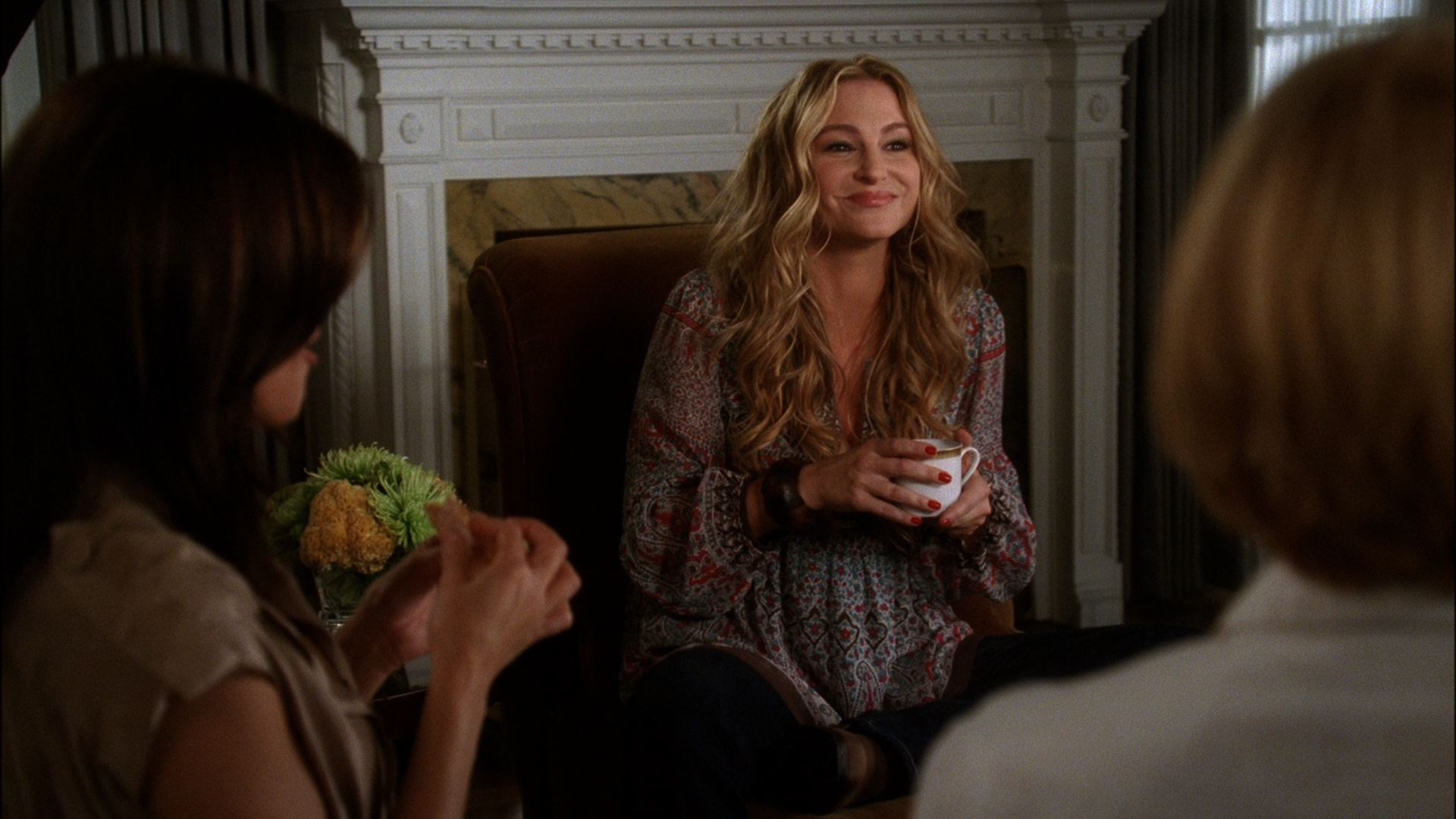 Angie Bolen (Drea de Matteo) sits smiling warmly while holding a teacup, flanked by two other women in the foreground whose backs and shoulders face the camera.