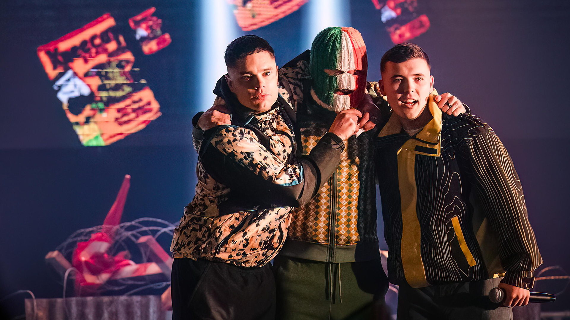 The image features the Irish hip-hop trio Kneecap, consisting of Mo Chara, Móglaí Bap, and DJ Próvai, known for their anarchic style and focus on the Irish language.