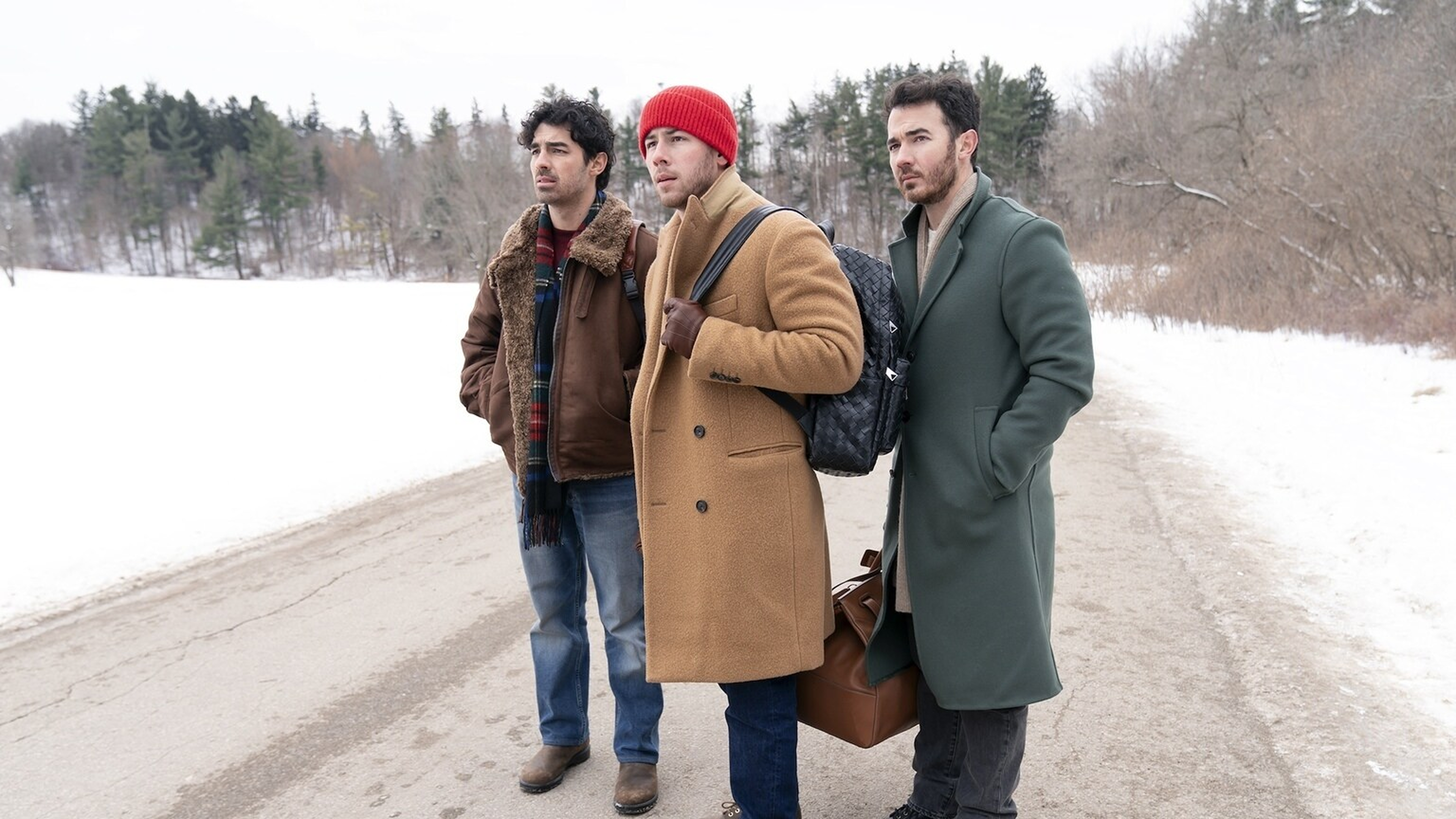 The Jonas Brothers stand in the middle of a country road covered in slush with fresh snow at the sides of the road.