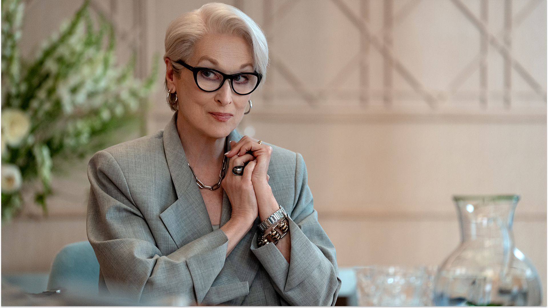 A promotional still from The Devil Wears Prada 2 shows Miranda Priestly (Meryl Streep) seated at a table, looking forward with a knowing smile and her hands clasped together. She sports her signature styled white hair, thick black-framed glasses, silver hoop earrings, a thick chain necklace, and a tailored grey oversized blazer.