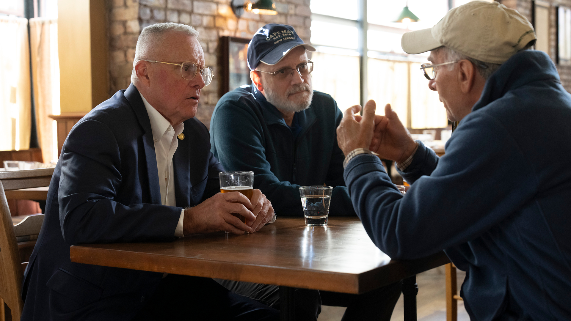 This image shows three men having a conversation at a wooden table in a pub setting, with one man in a suit wearing glasses and another in a cap.