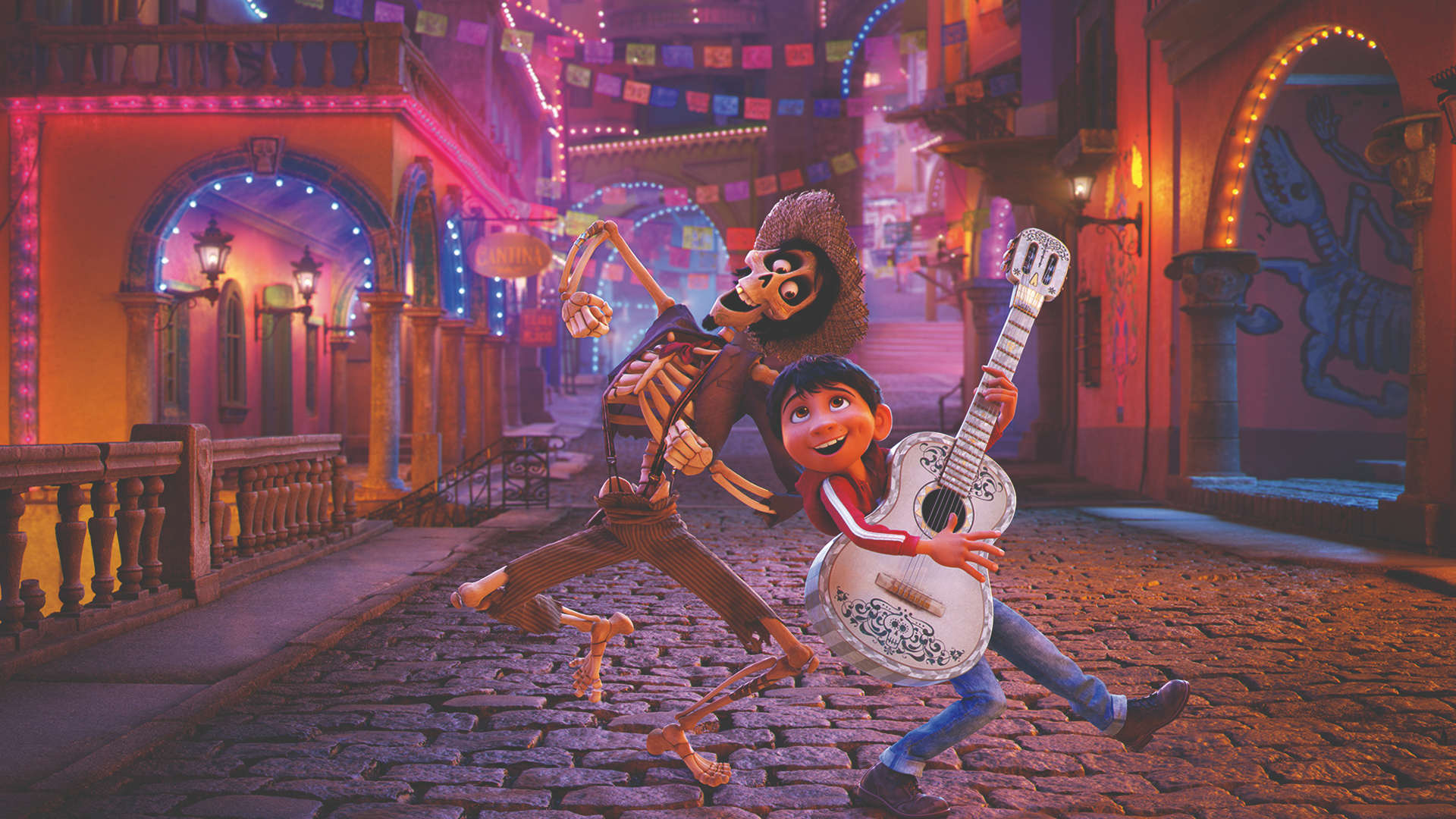 An animated, vibrantly lit street scene from Coco shows Miguel (Anthony Gonzalez) smiling while holding a white acoustic guitar next to Héctor (Gael García Bernal), a skeleton in ragged clothes wearing a straw hat, as they pose on a cobblestone street decorated with colorful festive banners and glowing lights.