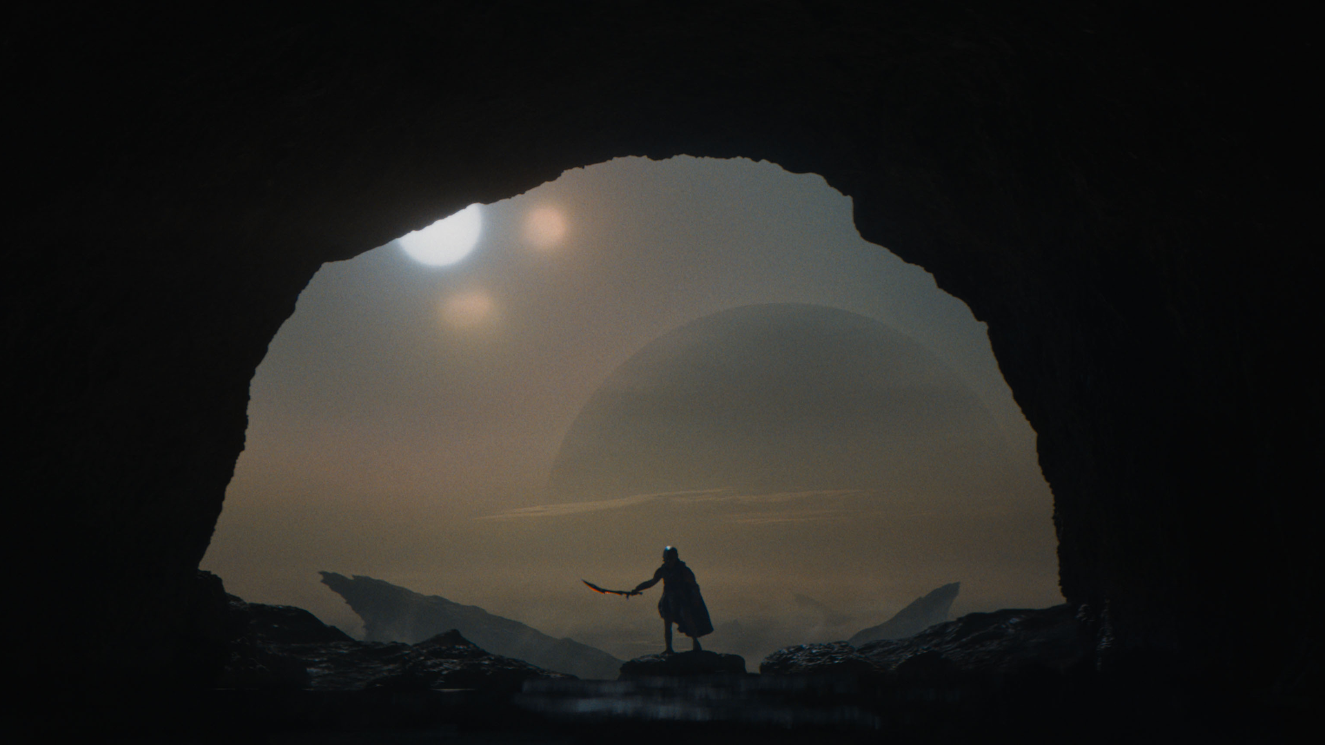 Dek (Dimitrius Schuster-Koloamatangi) stands at the mouth of a cave, wielding a weapon. A wide, barren landscape looms below.
