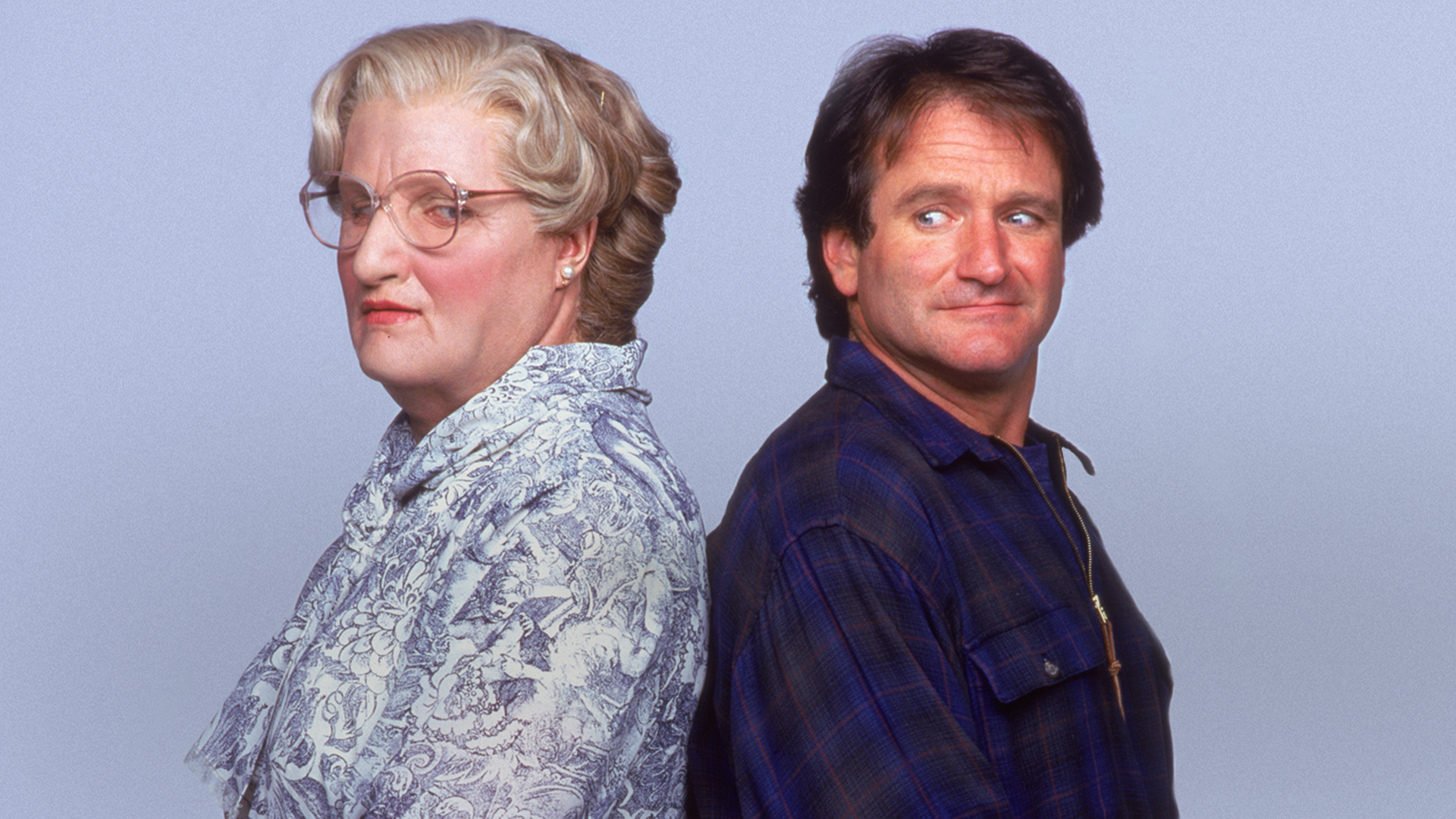 A classic promotional image from Mrs. Doubtfire shows Daniel Hillard (Robin Williams) standing back-to-back with his alter ego Mrs. Doubtfire (Robin Williams), with Daniel glancing over his shoulder with a subtle grin while Mrs. Doubtfire looks off to the side in a patterned blouse and glasses against a plain grey background.