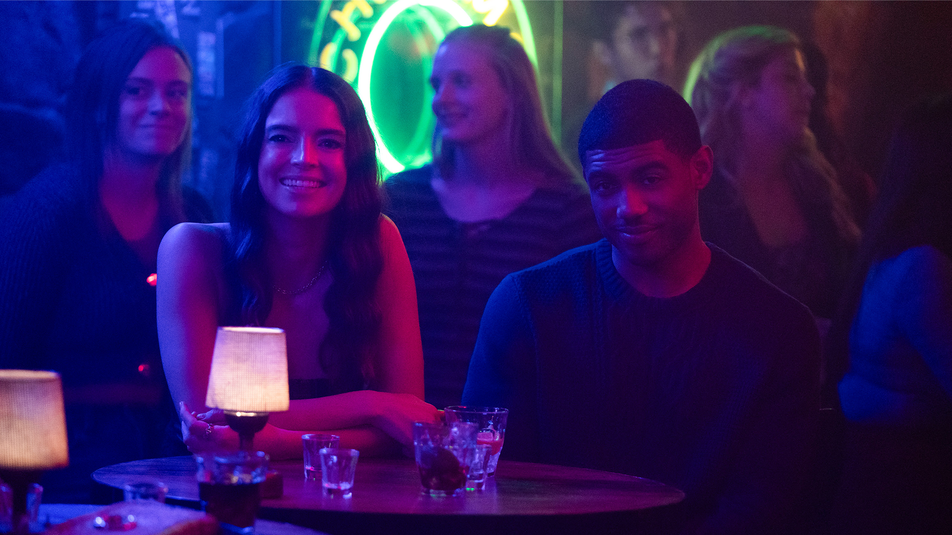 Two characters sit smiling at a table in a bar, covered in blue light.