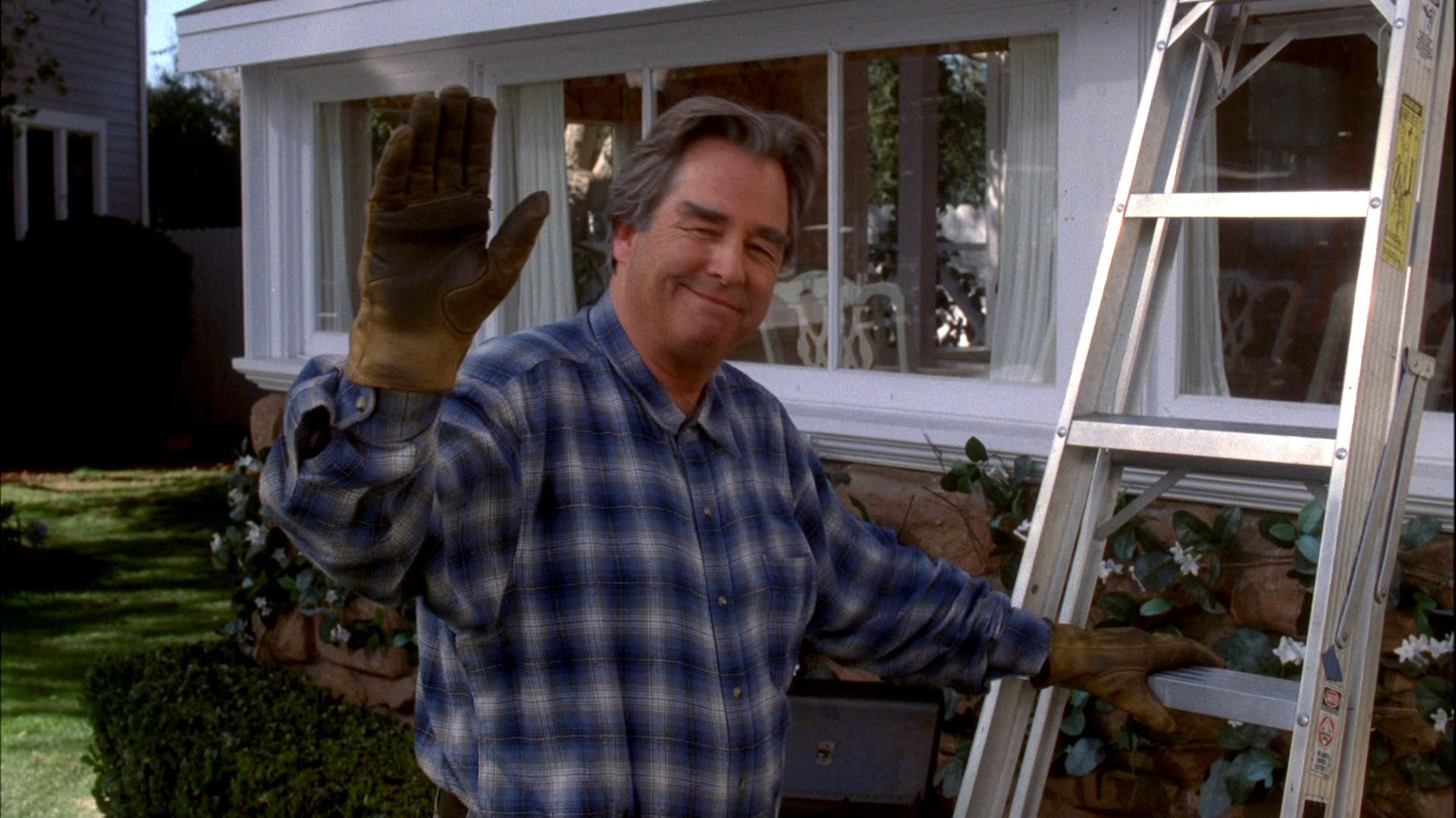 Eli Scruggs (Beau Bridges) smiles pleasantly and waves a gloved hand while standing outside in a yard next to an aluminum ladder.