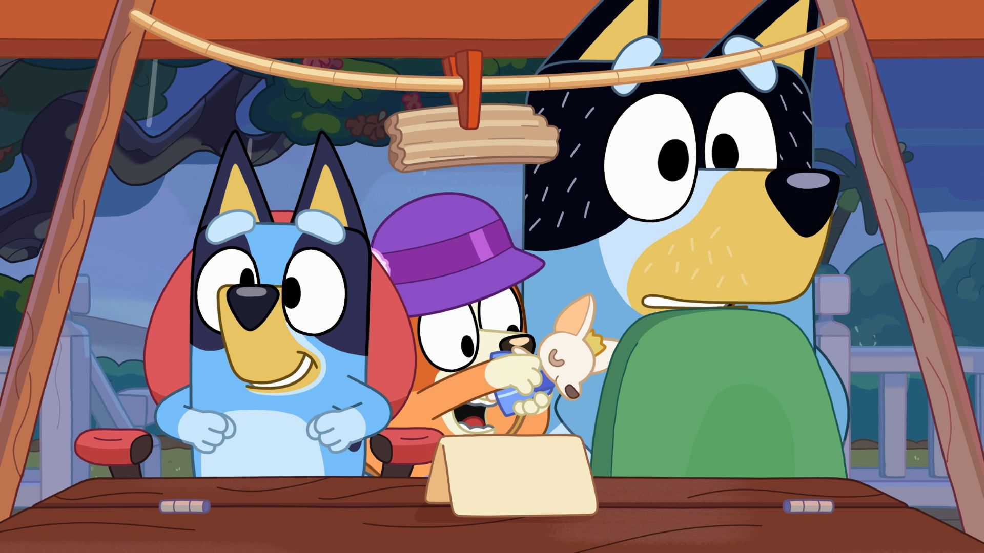 An animated scene from the show Bluey shows Bluey smiling enthusiastically next to Bingo, who wears a purple hat while holding a small toy dog, as Bandit looks on with wide eyes under a backyard tent structure.