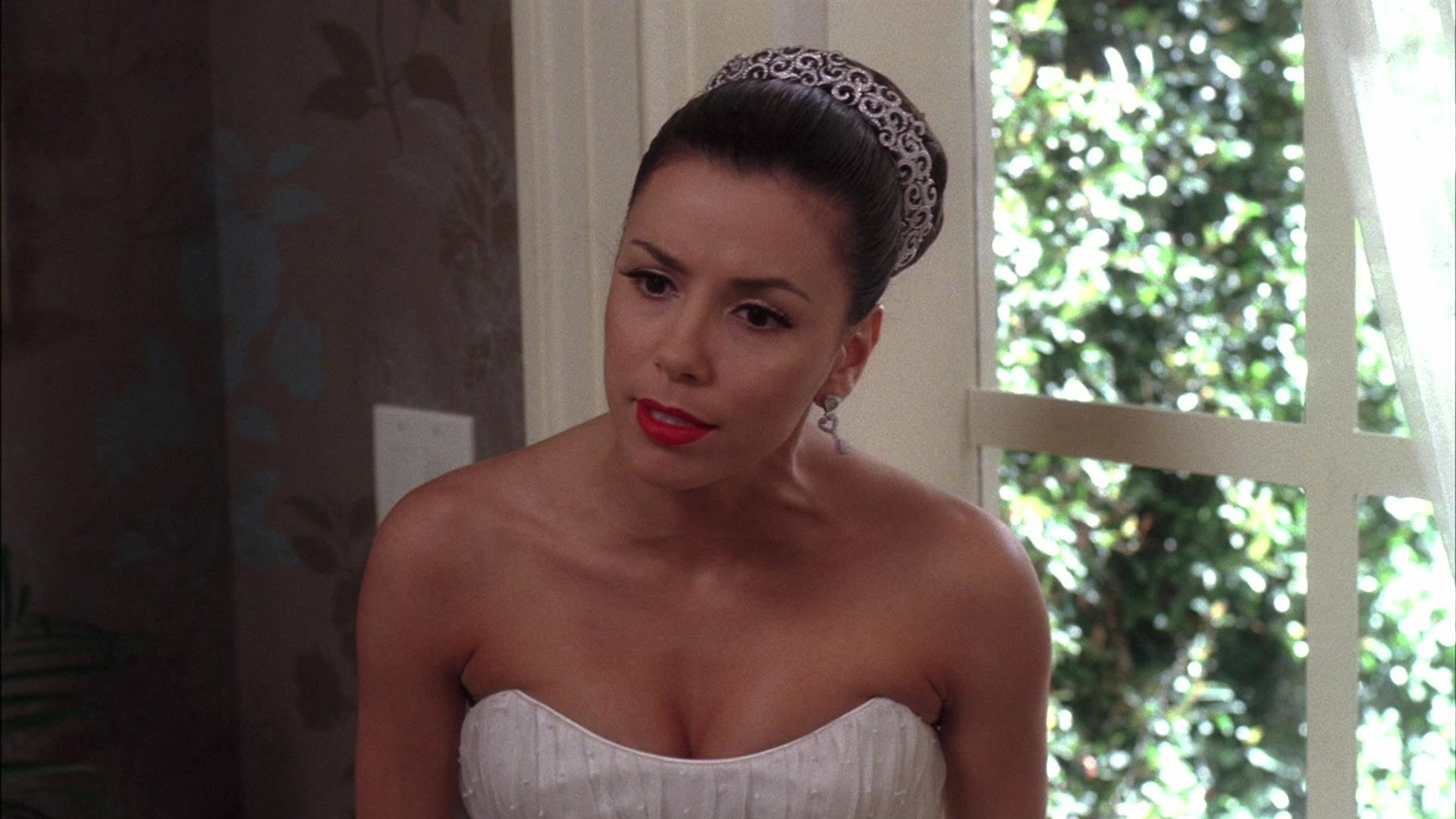 Gabrielle Solis (Eva Longoria) looks forward with a concerned expression, wearing a strapless white gown, a jeweled tiara over her updo, and bright red lipstick next to a bright window.