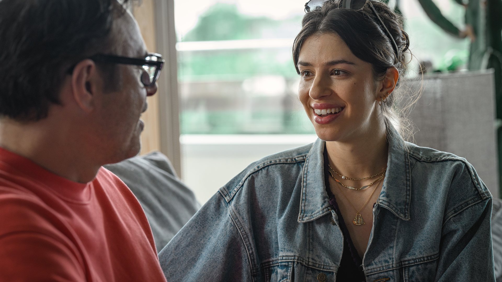 Izzy (Yali Topol Margalith) smiles brightly as she talks to Steve (Jemaine Clement), who is viewed from the side wearing glasses and a red shirt, in a scene from Alice and Steve.