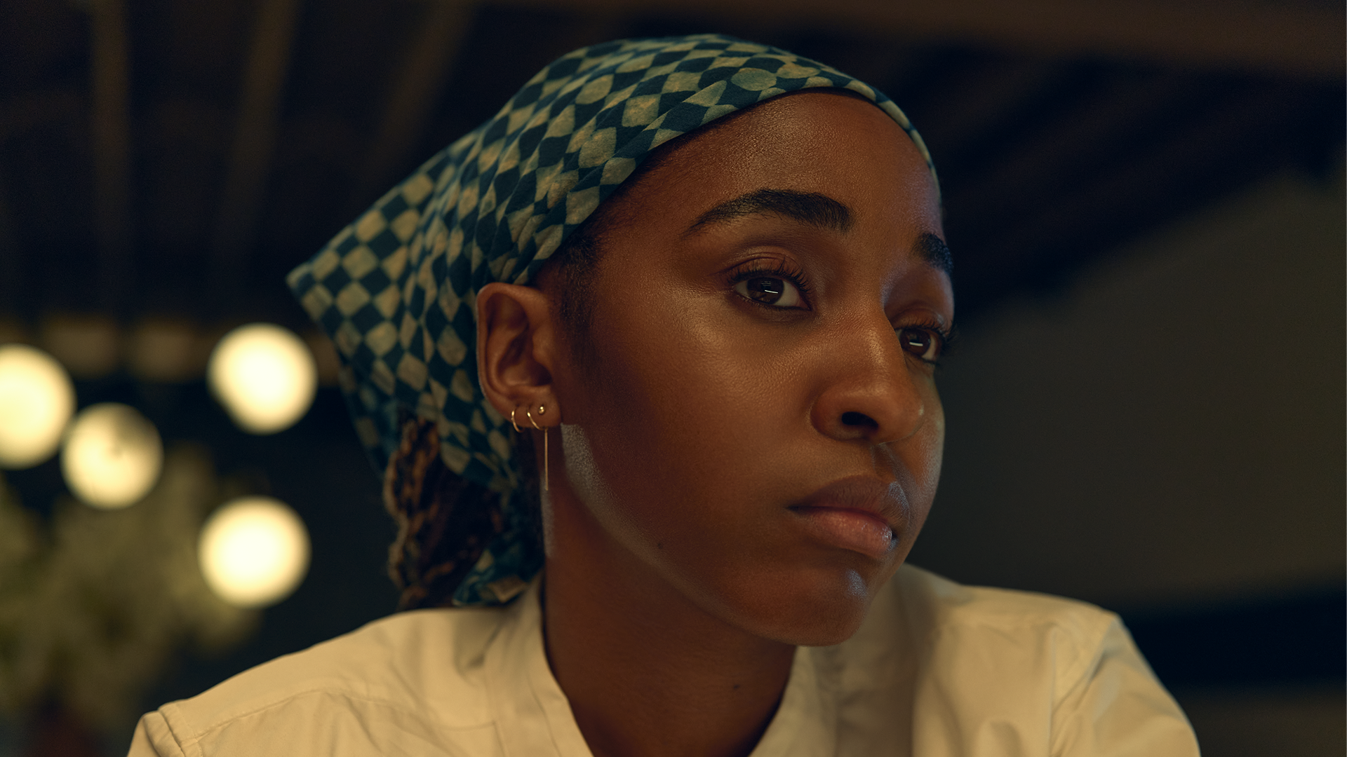 A close-up shot shows a focused Sydney Adamu (Ayo Edebiri) wearing a white chef's uniform and a green and white patterned headscarf while looking off to the side in a restaurant setting.
