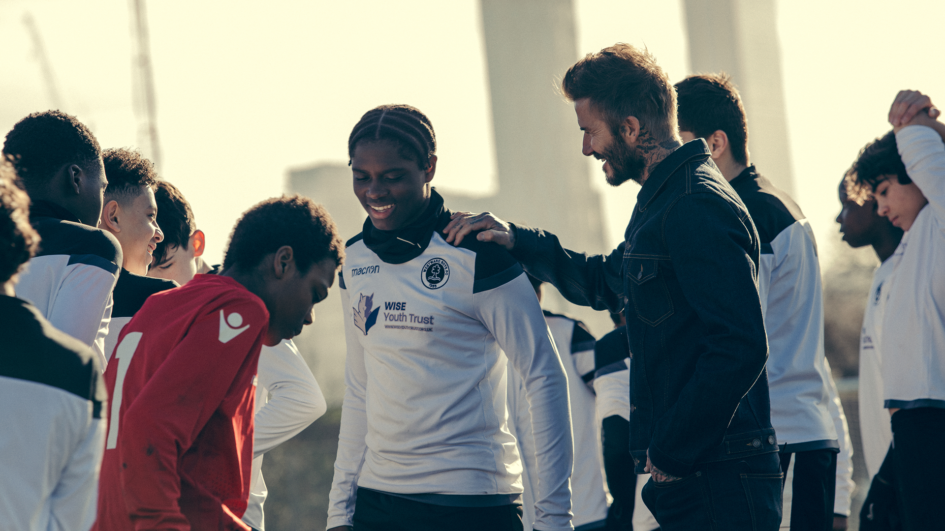 A sunny, outdoor scene from the sports docuseries Save Our Squad with David Beckham showing David Beckham in a denim jacket smiling and resting his hand on the shoulder of a smiling youth soccer player wearing a white Wise Youth Trust jersey on a training field.