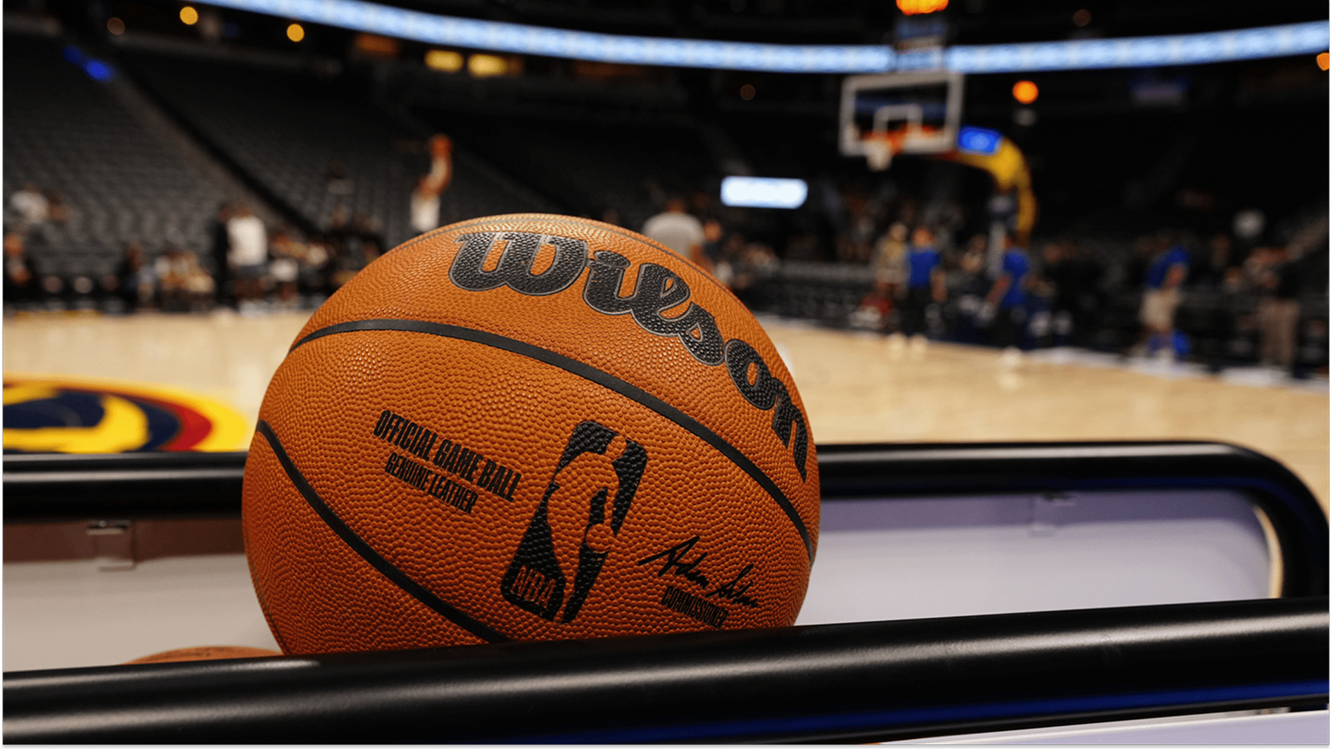 A close-up shot of an official Wilson NBA basketball resting on a rack, with a blurred basketball court and arena seats in the background.