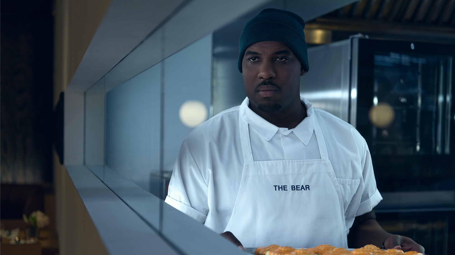 A medium shot of a serious Marcus Brooks (Lionel Boyce) wearing a blue beanie and a white chef's apron embroidered with "THE BEAR," looking off-camera while holding a tray of baked goods in a professional kitchen.