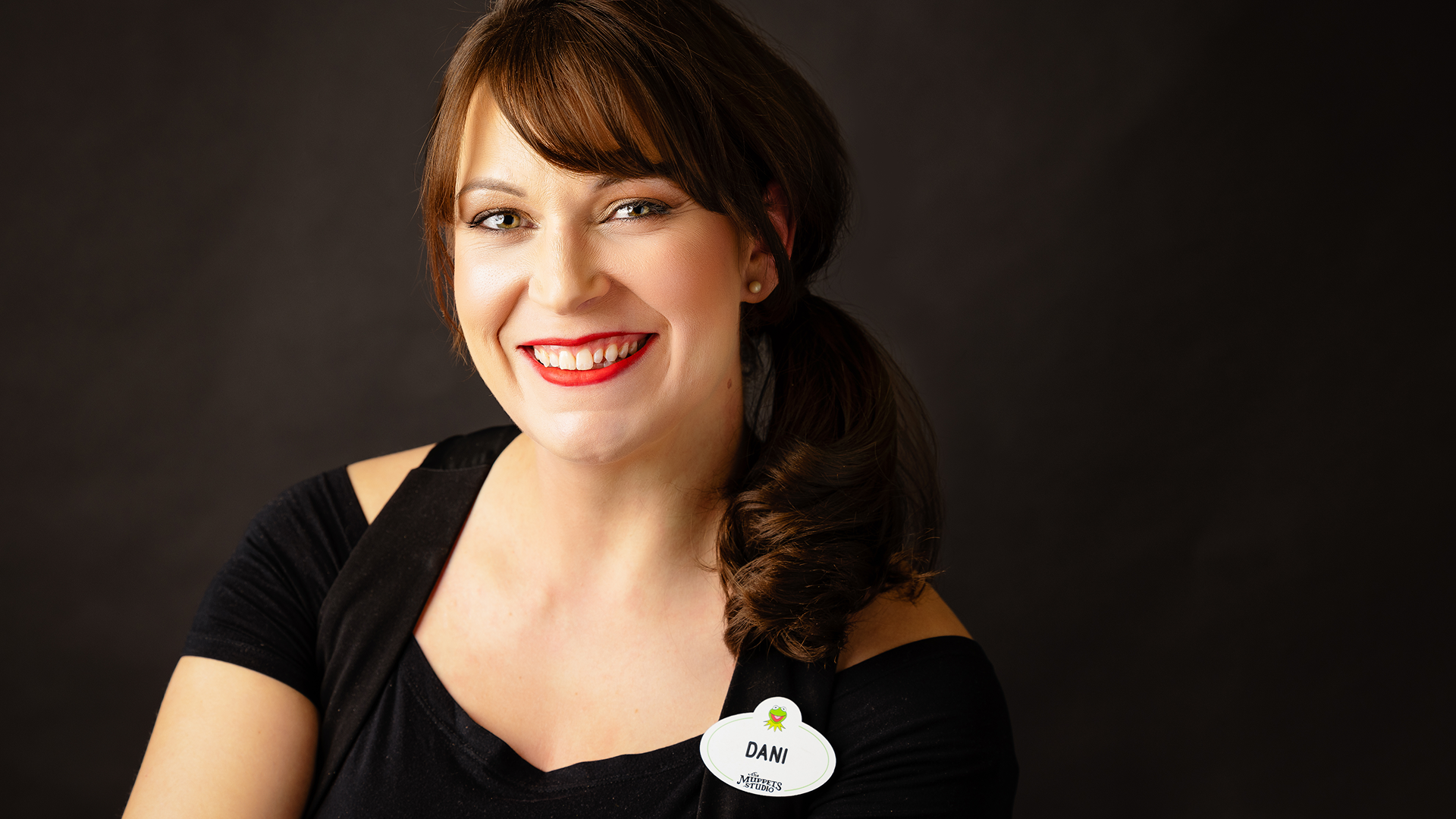 A headshot of Dani Iglesias, Creative Director of The Muppets Studio, smiling in front of a dark background and wearing a Muppets Studio employee name tag.