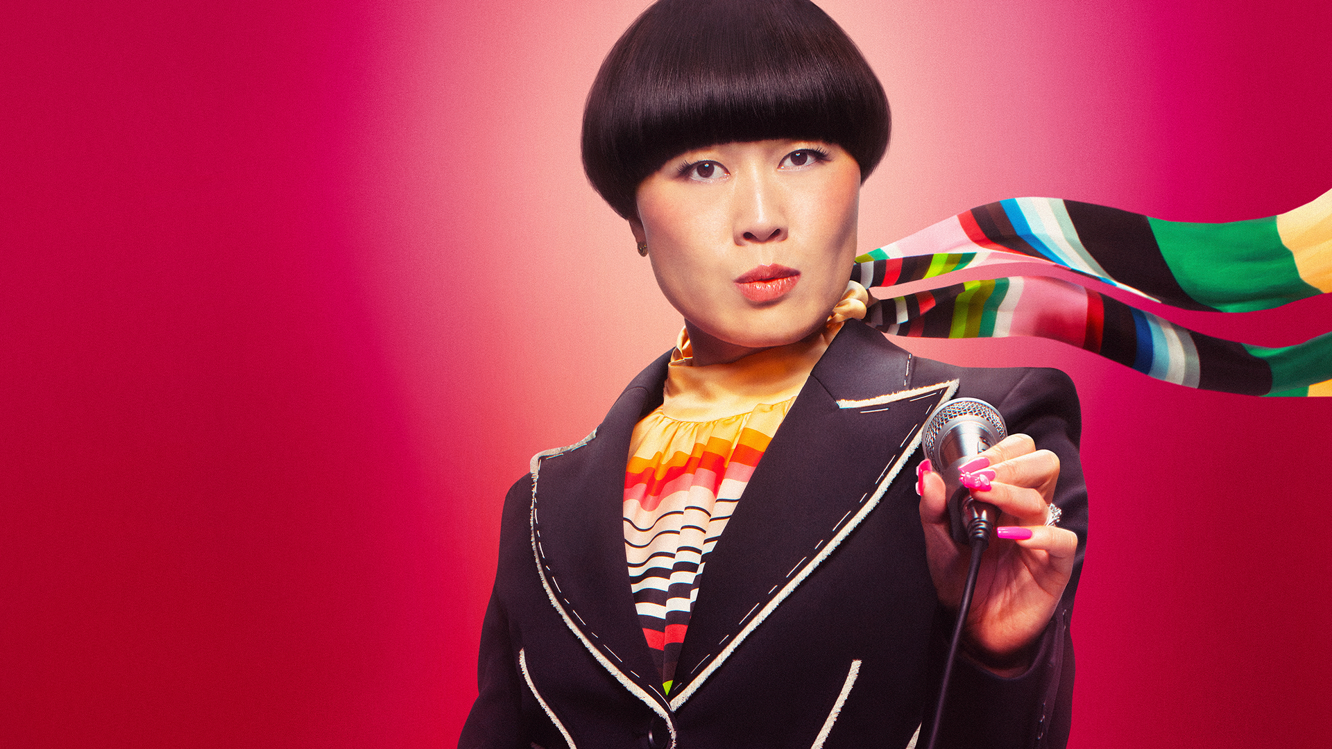 Atsuko Okatsuka looks into the camera holding a microphone in front of a red background