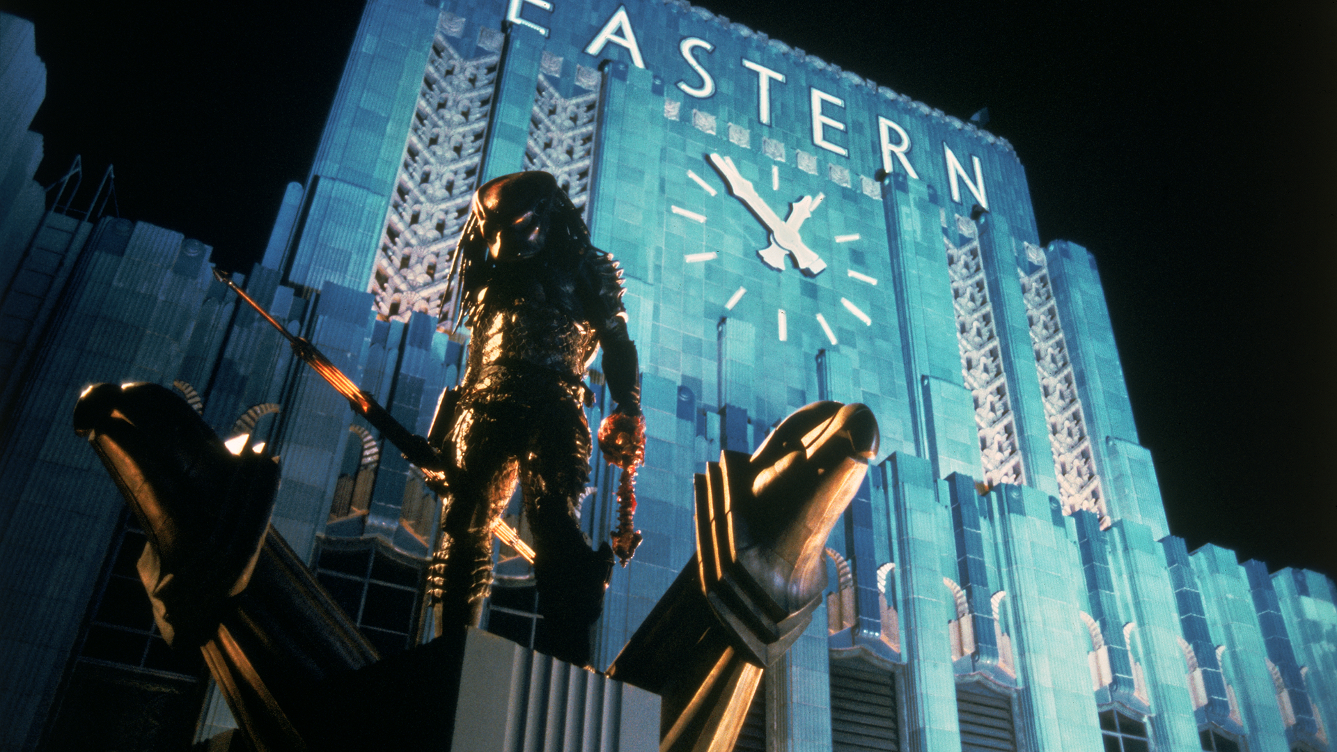 El Depredador (Kevin Peter Hall) está de pie en lo alto de un edificio por la noche, con una torre de reloj en el fondo