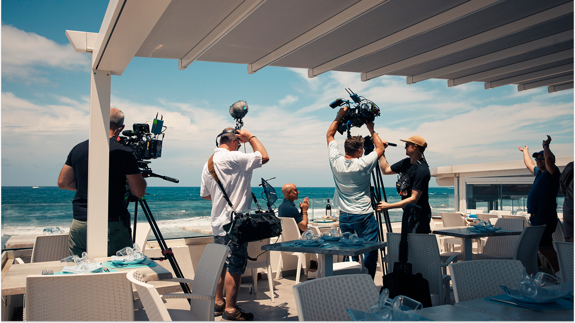 A camera crew films Stanley Tucci sat at a dining table in the outside area of a restaurant by the sea