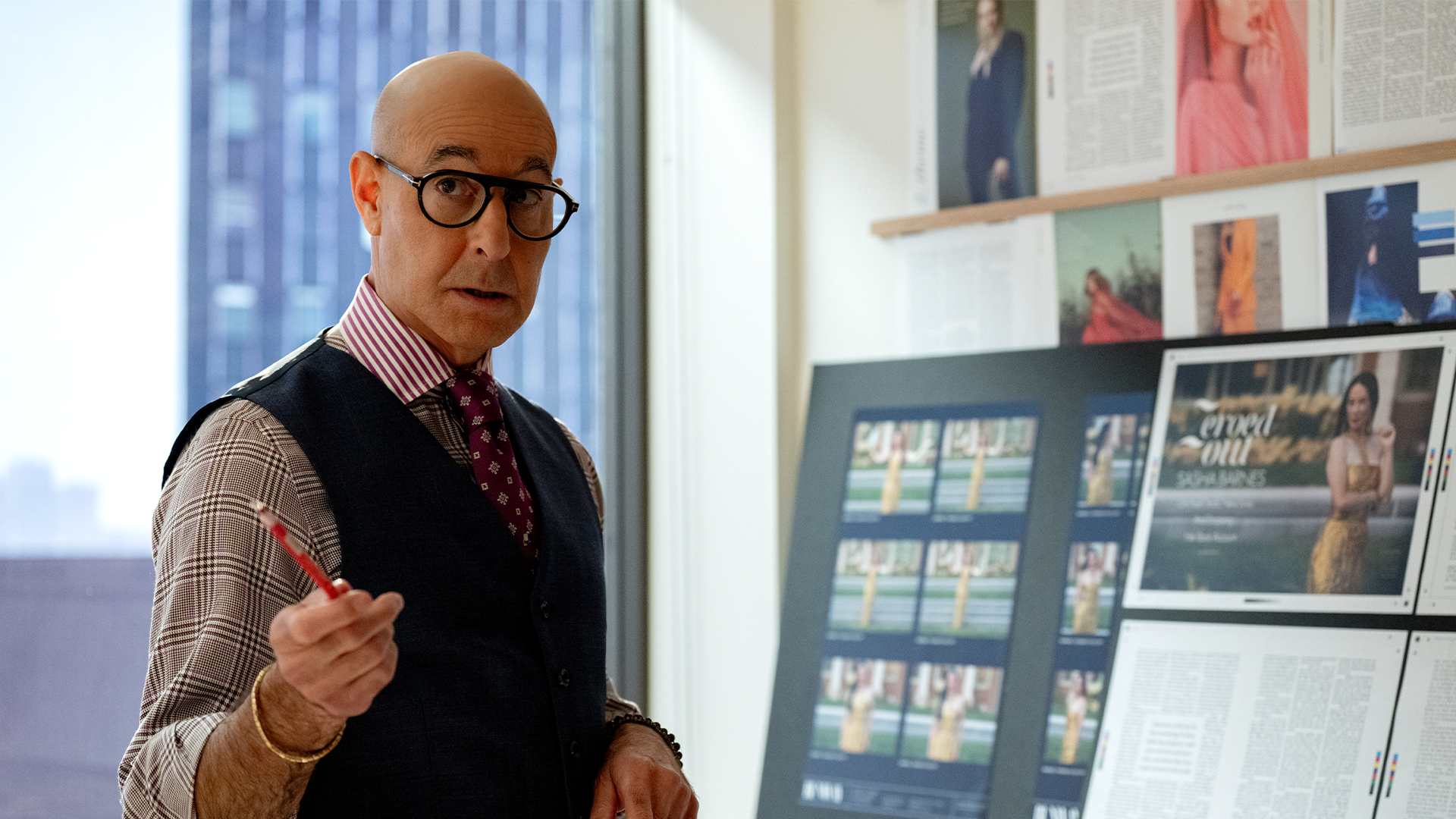 A promotional still from The Devil Wears Prada 2 shows Nigel Kipling (Stanley Tucci) standing in a brightly lit fashion office, holding a red pencil and gesturing while speaking with a surprised expression. He wears thick round black glasses, a navy waistcoat over a brown houndstooth shirt, a red-and-white striped collar, and a patterned purple tie, with fashion layout boards visible behind him.