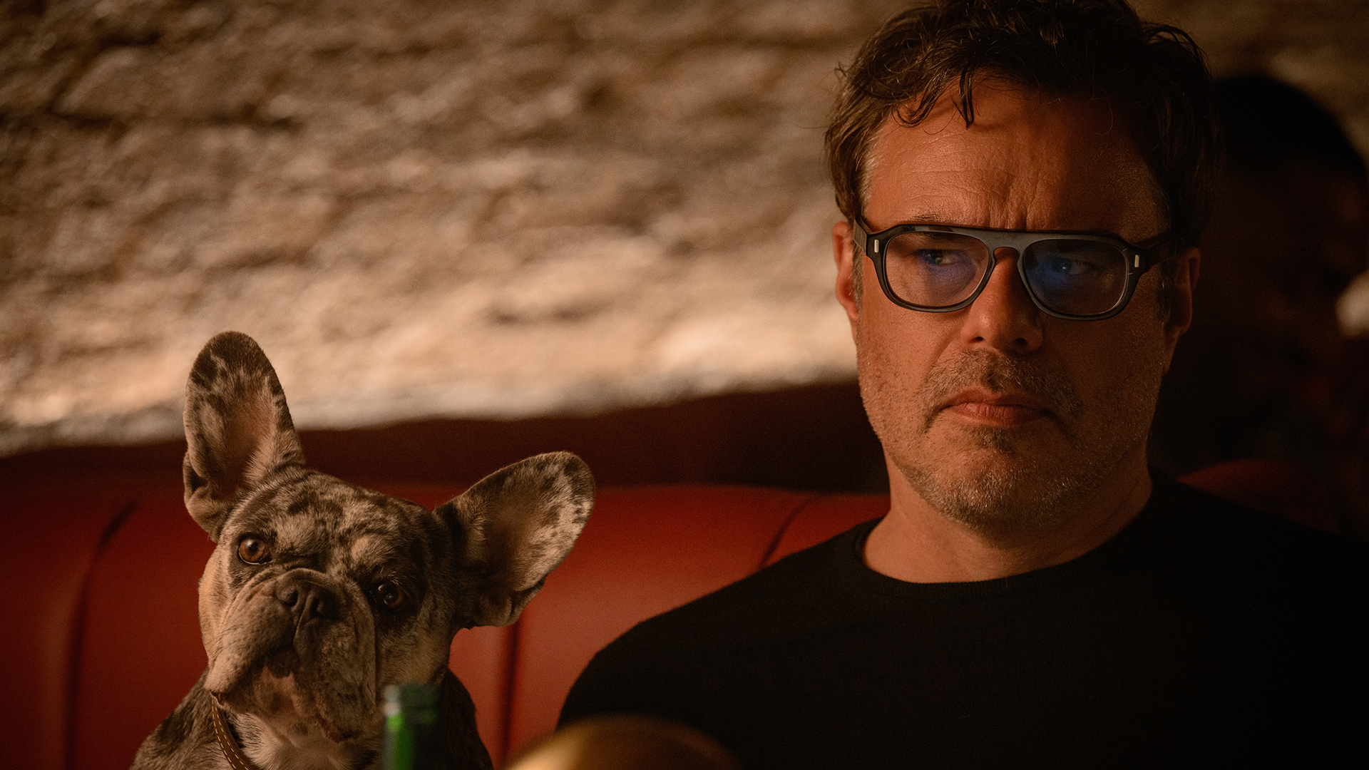 Steve (Jemaine Clement) sits with a serious expression next to a brindle French Bulldog on a red leather booth in a dimly lit scene from Alice and Steve.