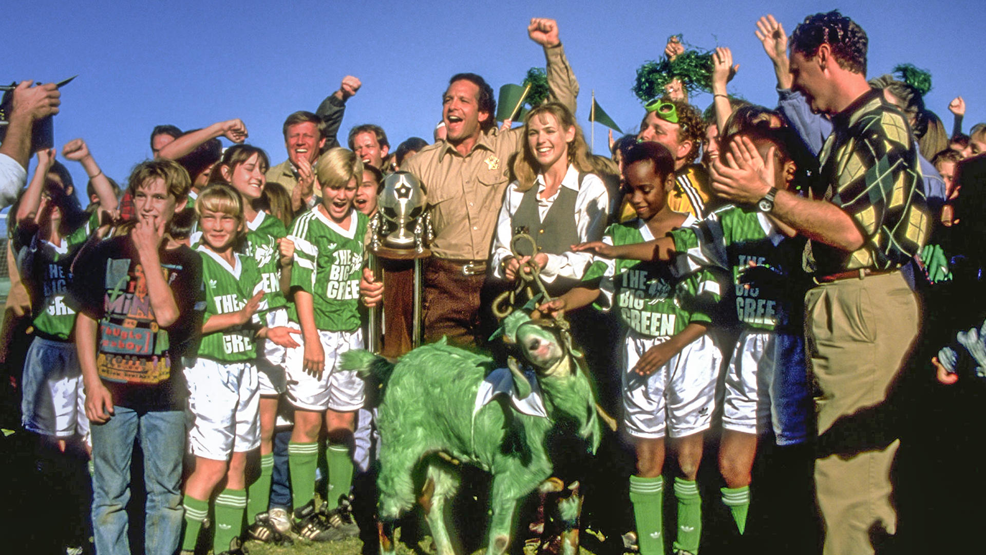 A celebratory scene from the sports comedy film The Big Green showing Sheriff Tom Palmer (Steve Guttenberg) and Miss Anna Montgomery (Olivia d'Abo) cheering with a gold trophy alongside their youth soccer team, who are gathered around a live goat painted completely green.