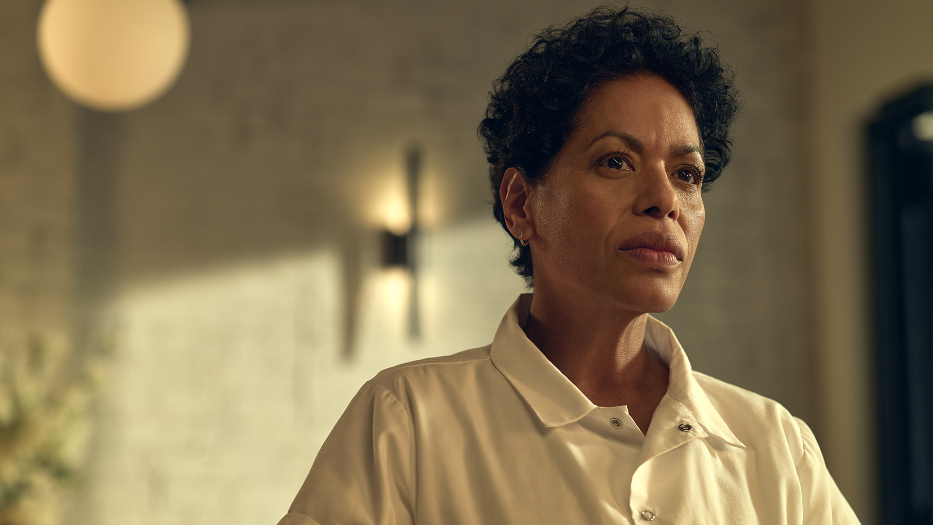 A medium close-up shot of a determined Tina Marrero (Liza Colón-Zayas) with short, curly dark hair, wearing a white chef's shirt and looking intently off-camera inside a brightly lit restaurant.