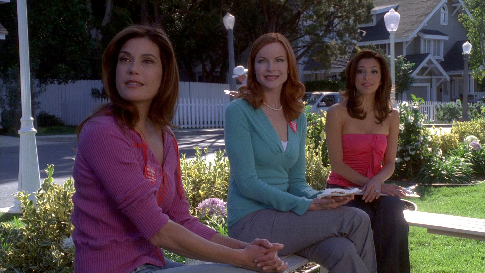 A scene from "Desperate Housewives" showing Susan, Bree, and Gabrielle sitting outdoors on a suburban lawn, smiling as they look off-camera.