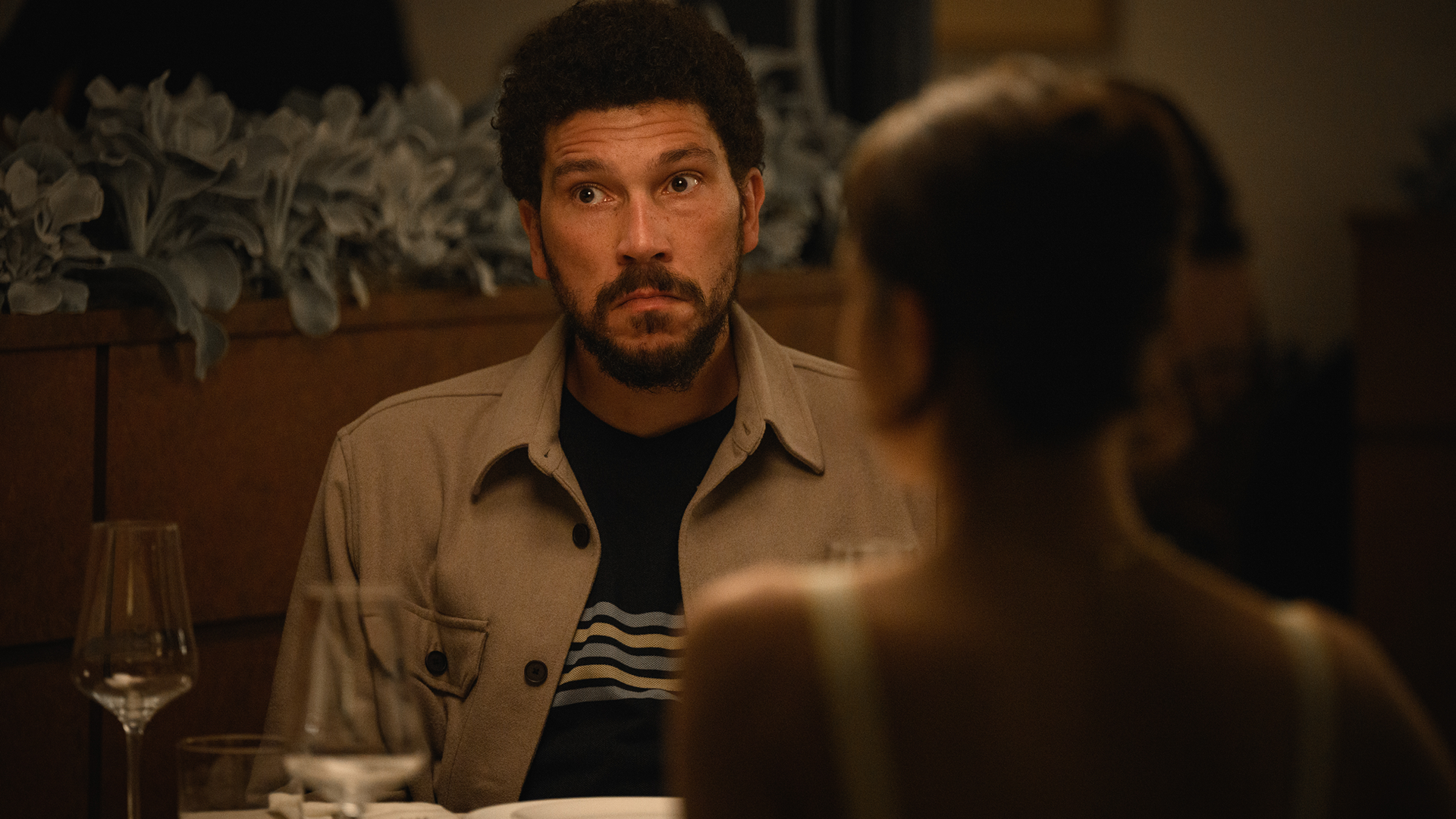 Daniel (Joel Fry) sits at a dinner table looking startled and wide-eyed across from a woman seen from behind in a scene from the comedy-drama series Alice and Steve.